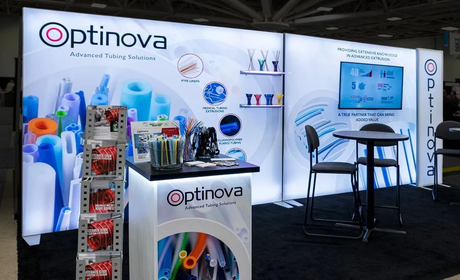 Optinova exhibit with colorful tubing graphics, sample products, a white branded counter and a wall-mounted screen.