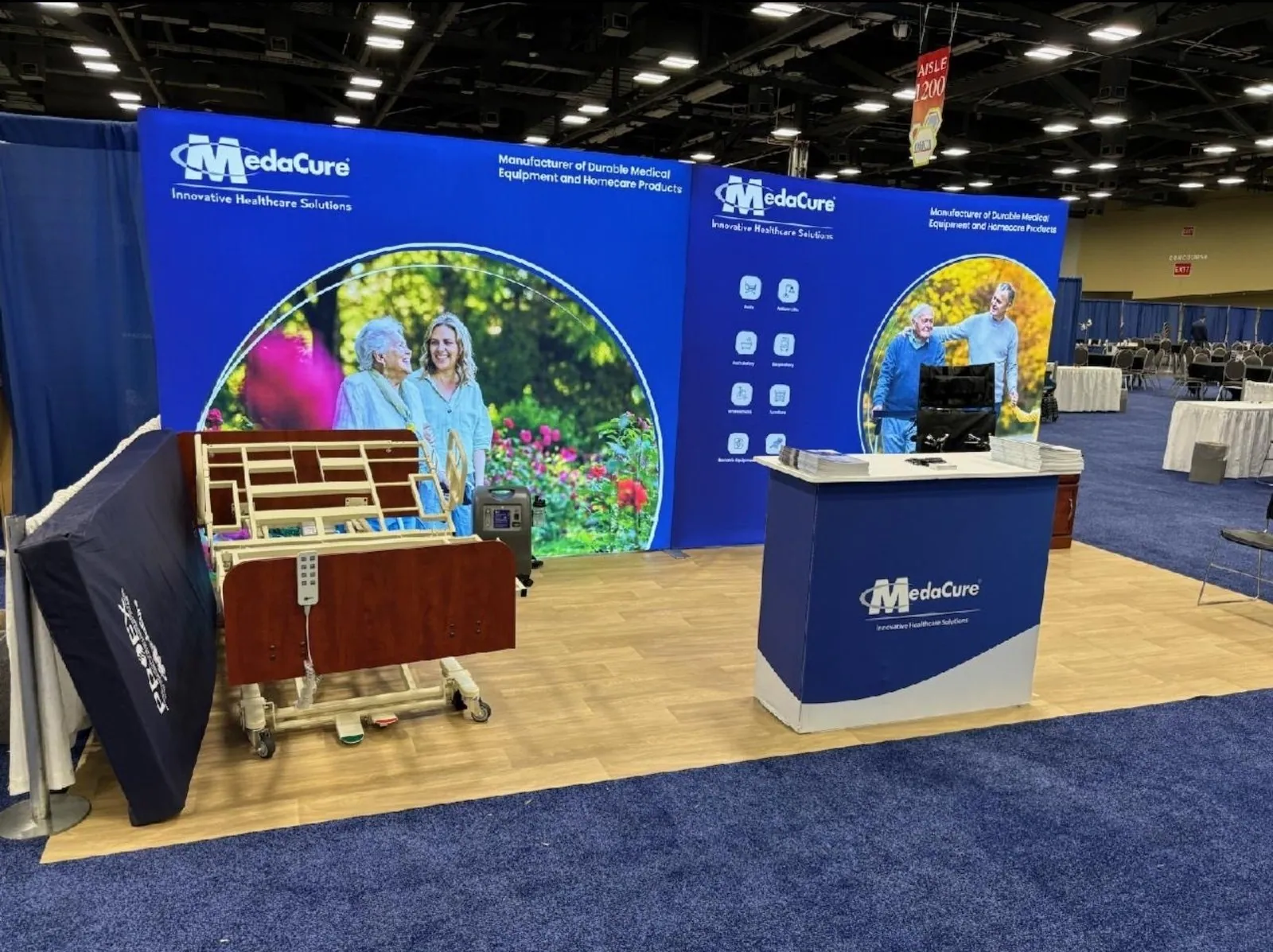 MedaCure exhibit with a hospital-bed demonstration, blue healthcare graphics, a reception counter and a monitor.