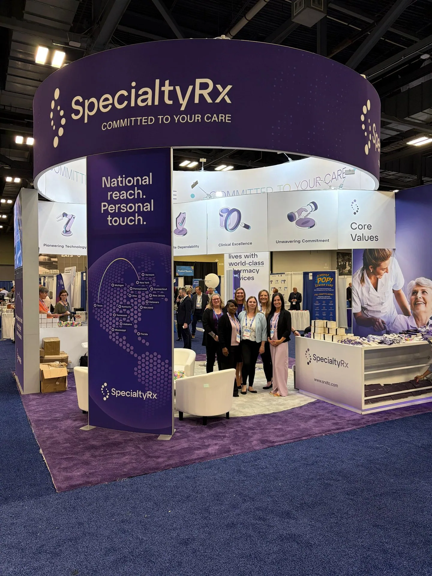 SpecialtyRx exhibit with a large curved purple overhead sign, white lounge seating, a branded counter and team members.