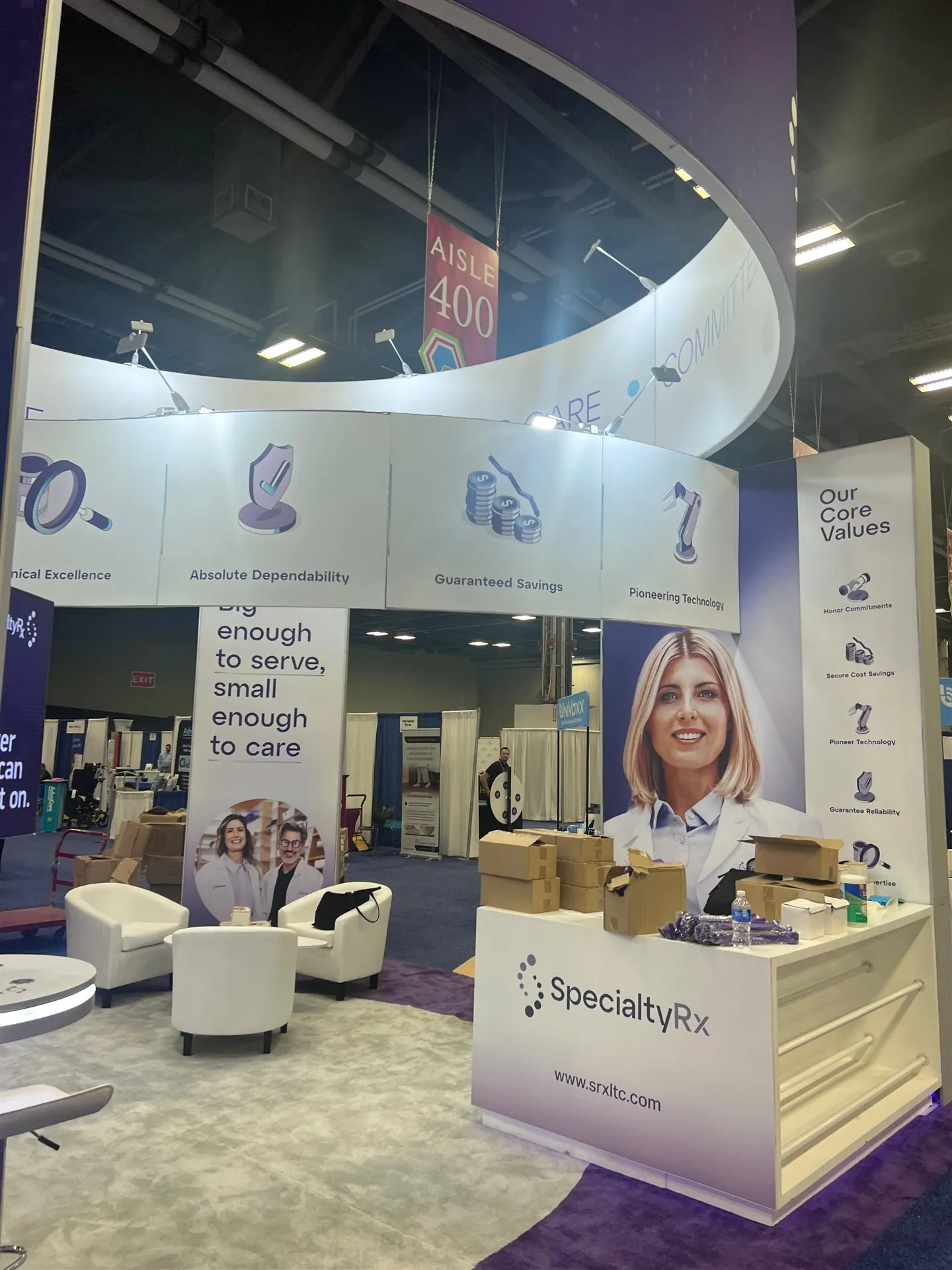 SpecialtyRx exhibit with a white reception counter, pharmacy messaging, purple accents and white lounge chairs beneath a curved canopy.