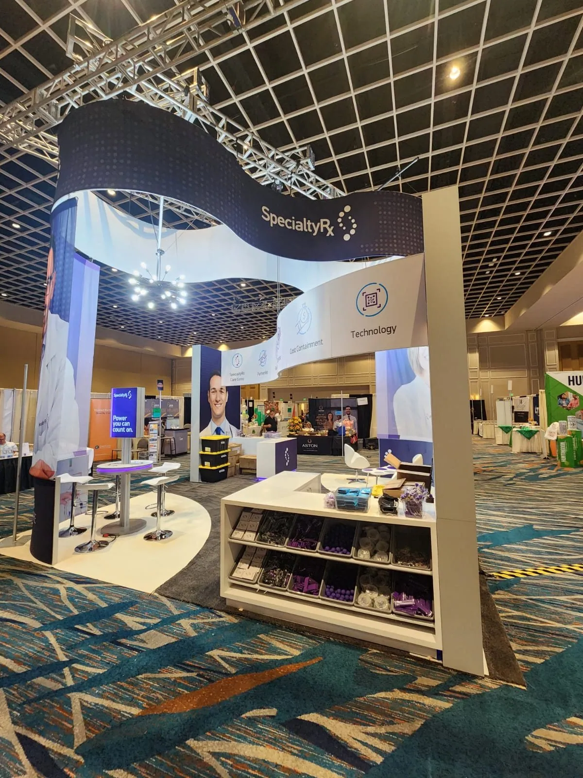 SpecialtyRx pavilion with a curved overhead canopy, hanging lights, consultation tables and a branded product counter.