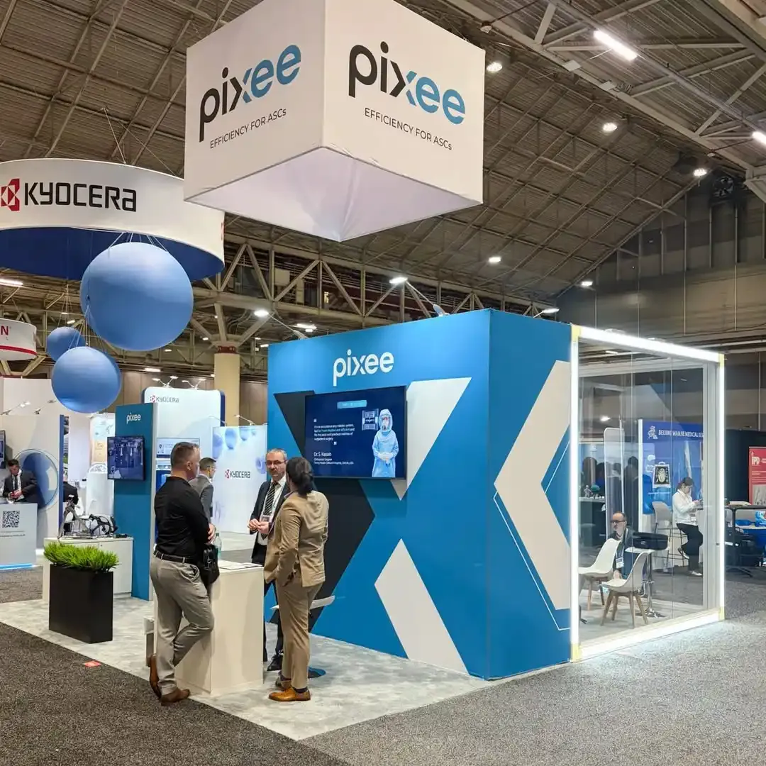 Visitors talking at Pixee Medical's blue exhibit beside a presentation screen and glass meeting room beneath a white hanging sign.
