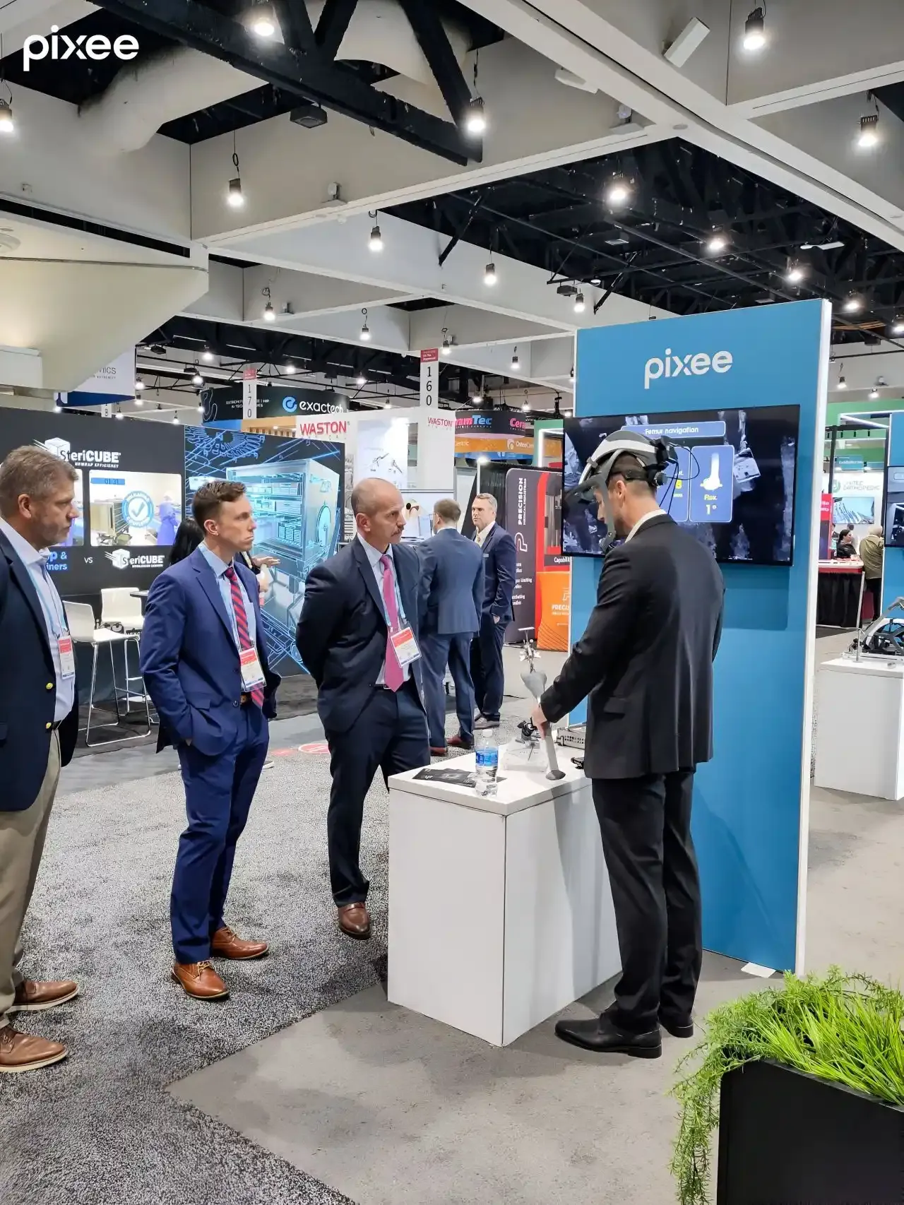A Pixee Medical presenter wearing an augmented-reality headset demonstrates surgical technology to visitors beside a branded counter and screen.