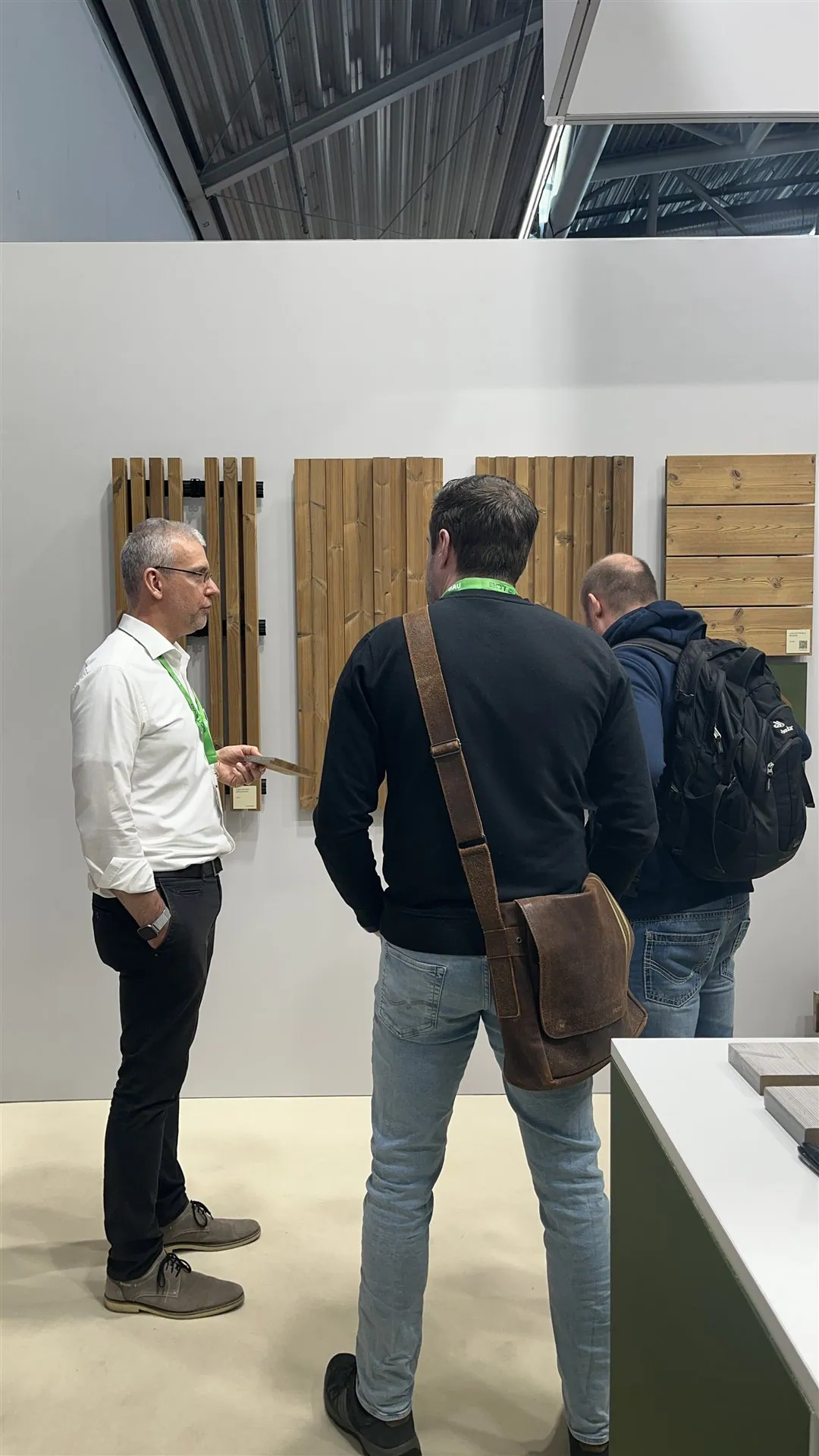 Three visitors talking beside a wall of vertical timber samples in Lunawood’s white exhibit.