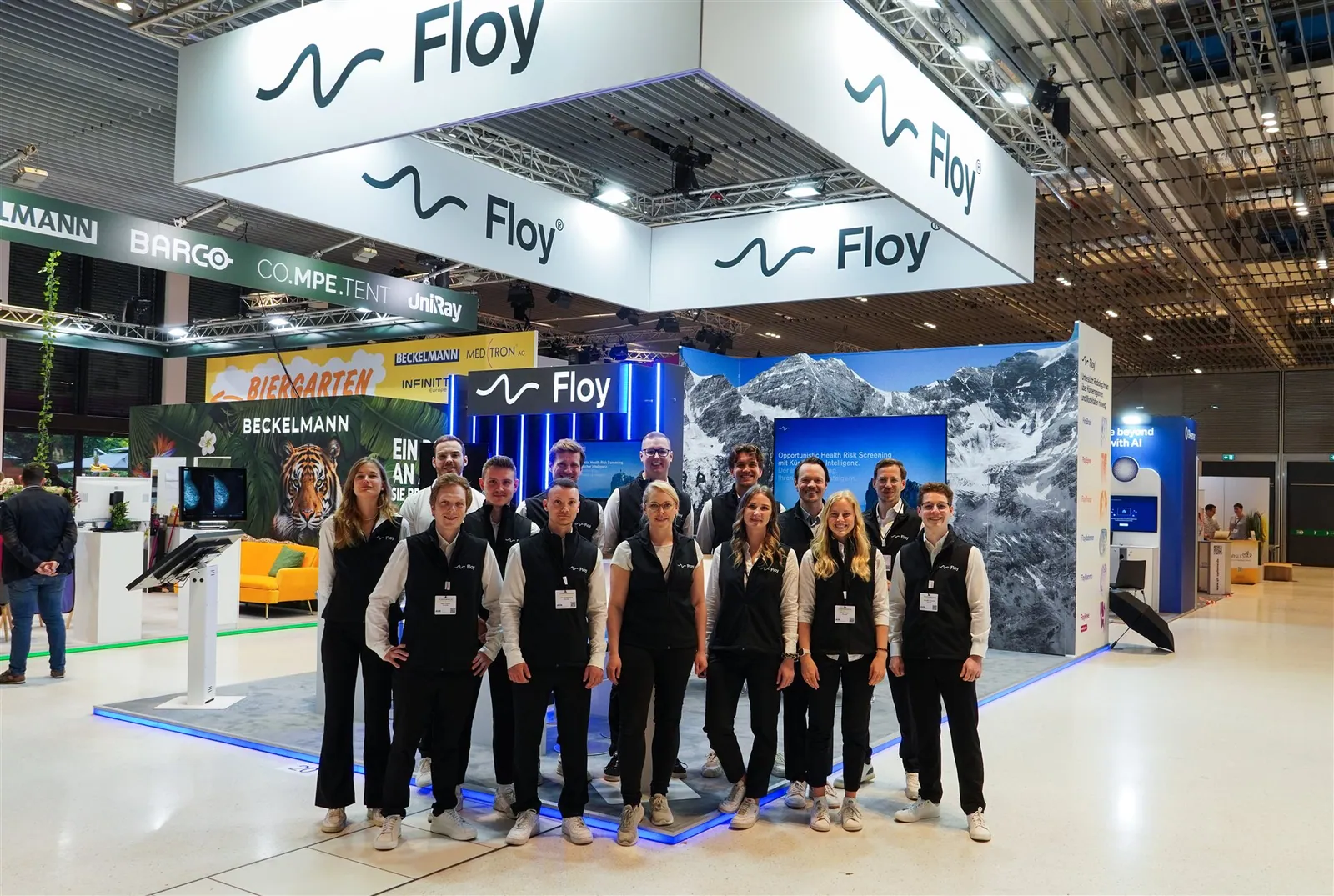 Floy team posing in a medical-technology exhibit beneath large suspended white signs, with imaging workstations and mountain graphics behind them.
