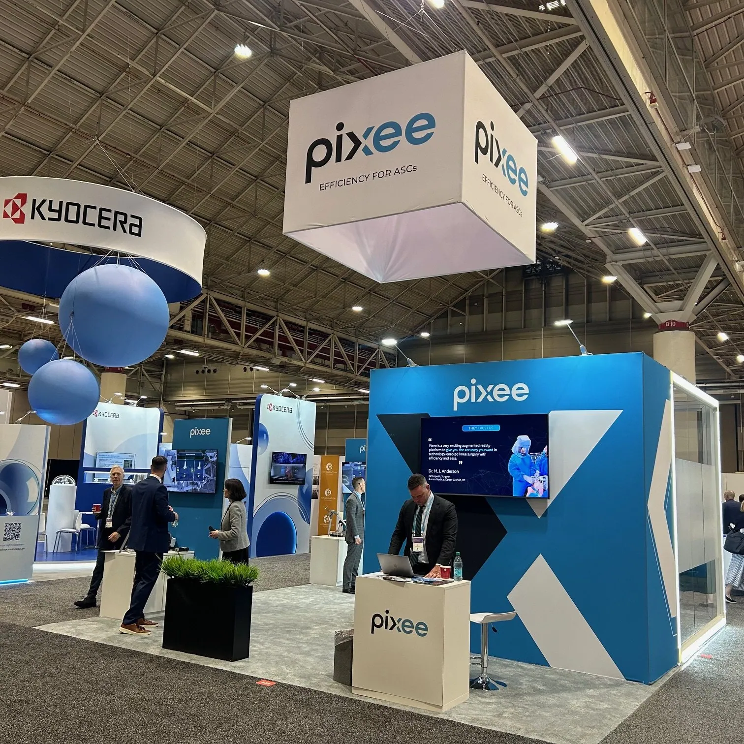 Pixee Medical exhibit with blue product walls, surgical-technology screens, visitors and a large white hanging cube sign.