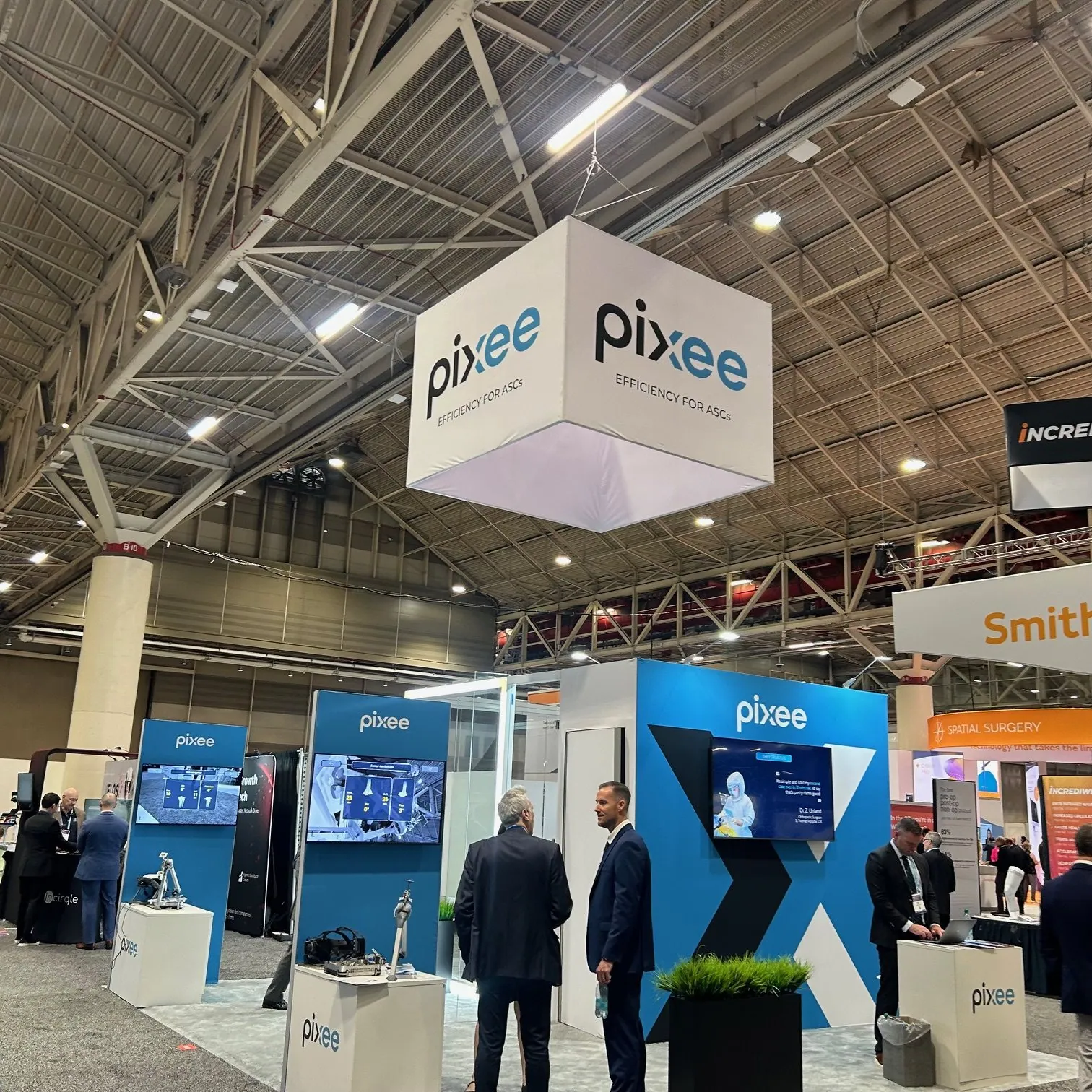 Visitors talking beside Pixee Medical's blue demonstration walls, surgical models and screens beneath a suspended cube-shaped sign.
