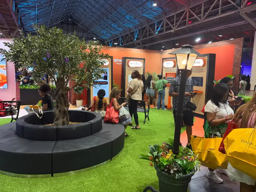 Visitors relaxing on curved seating around a tree inside Amazon’s activation, with artificial grass and orange storefront graphics.