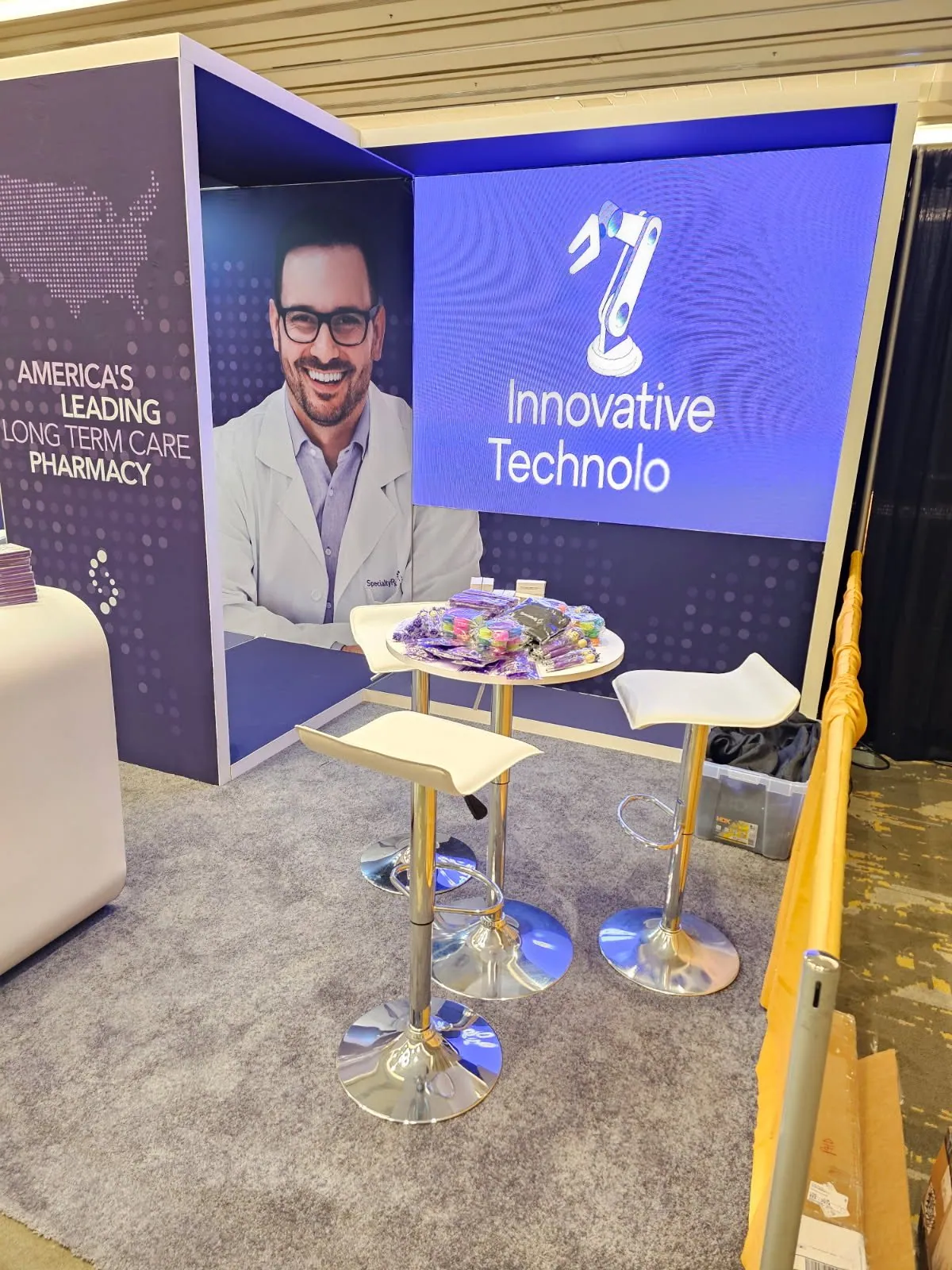SpecialtyRx exhibit corner with purple technology messaging, a large display screen and a small table with white stools.