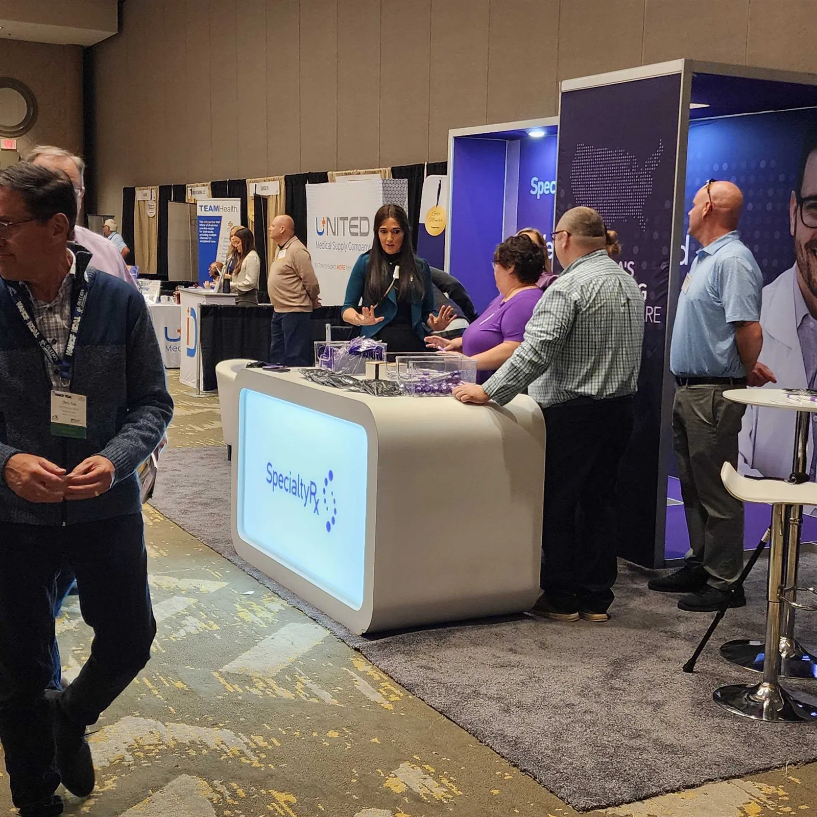 Visitors talking with the SpecialtyRx team around an illuminated white reception counter in a purple pharmacy exhibit.