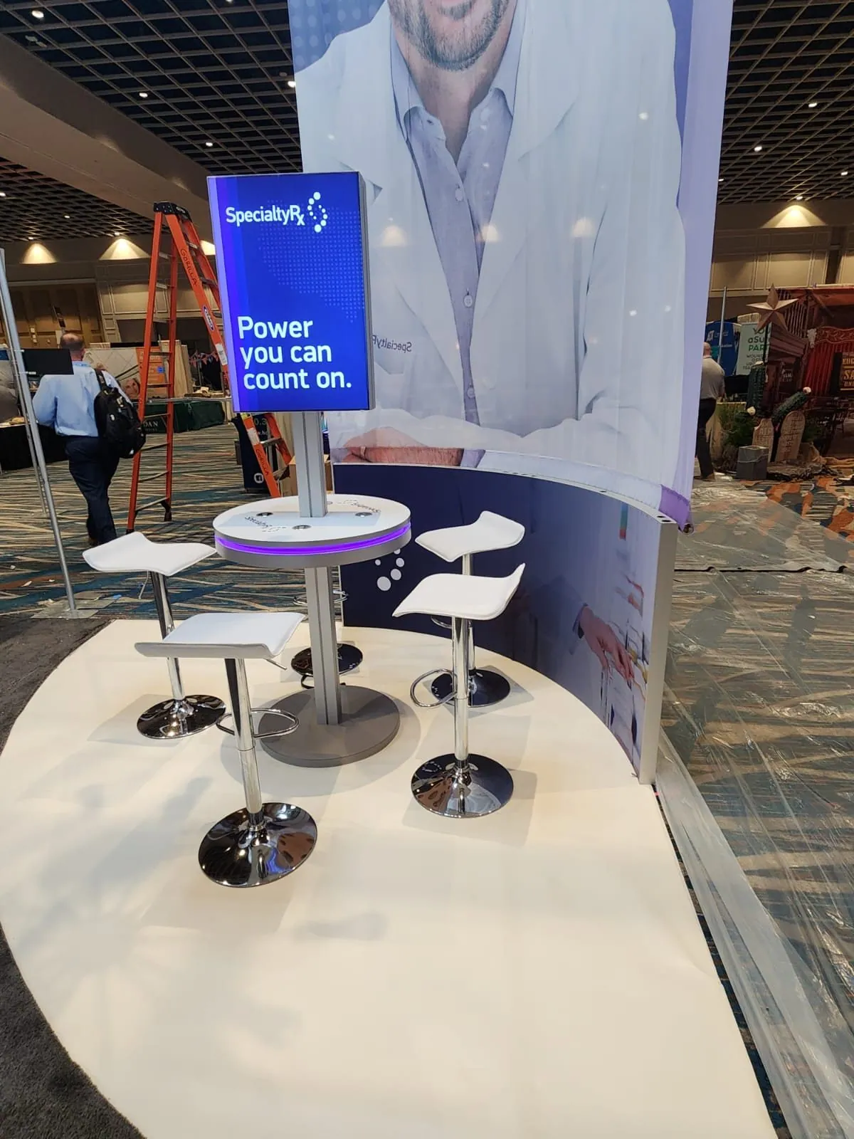 SpecialtyRx exhibit detail with a circular white table, two chairs and a vertical purple branded panel beneath a curved canopy.