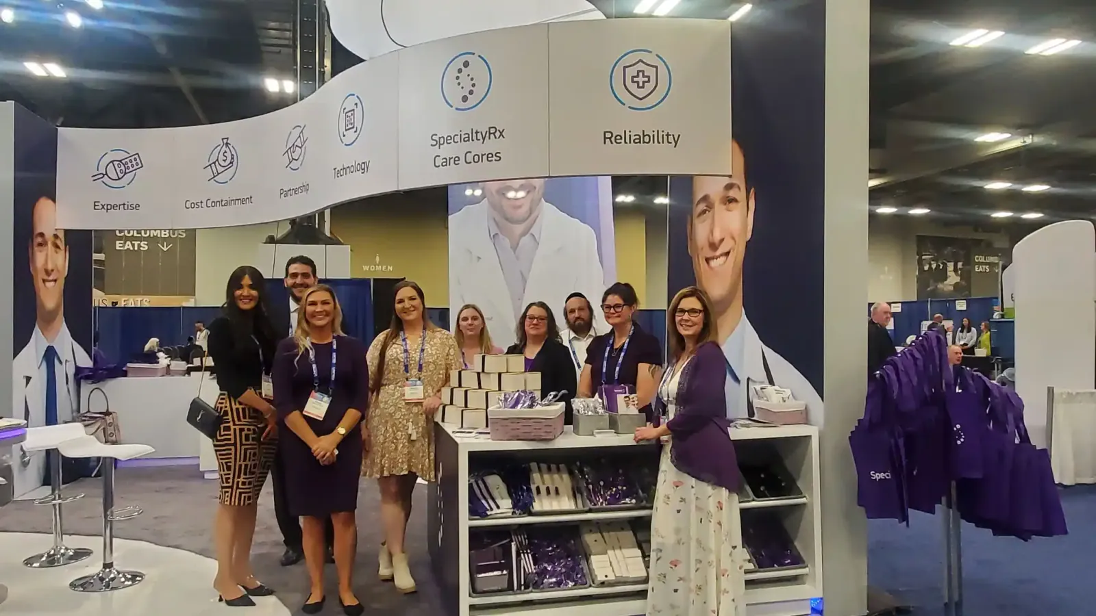 People posing at SpecialtyRx's exhibit around a giveaway counter, beneath a curved canopy listing pharmacy-service commitments.