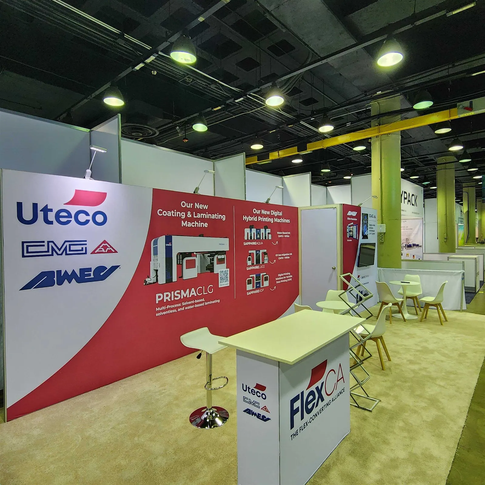FlexCA printing and converting exhibit with Uteco, CMG and Bimec wall graphics, a branded counter and white meeting tables.