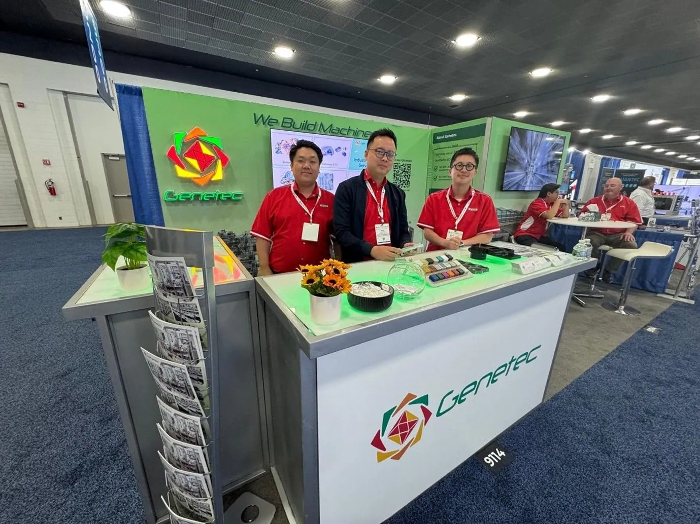 Three people posing behind Genetec Technology's illuminated counter, with component samples, green manufacturing graphics and screens behind them.