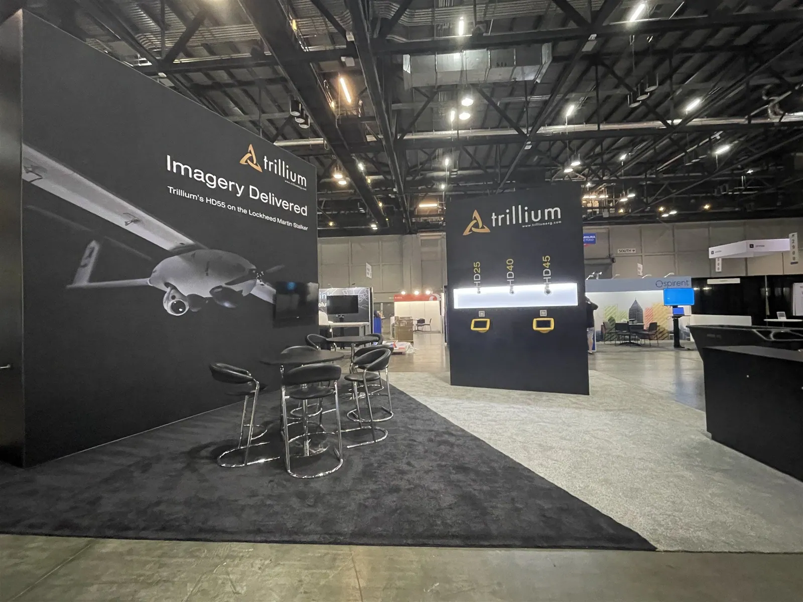 Trillium Engineering exhibit with dark aircraft graphics, illuminated camera-sensor displays, a reception counter and high meeting tables.