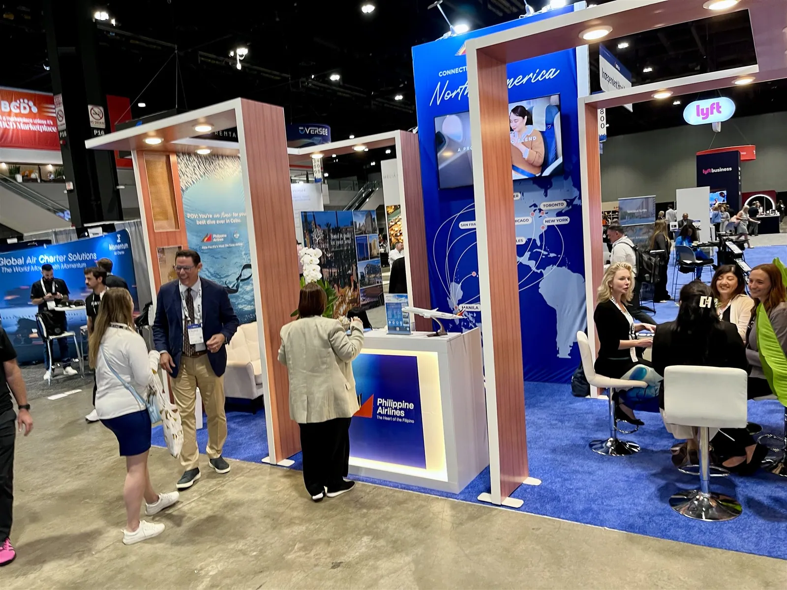 Visitors talking beside Philippine Airlines’ illuminated counter and meeting tables, with a blue North America route-map tower and warm framed walls behind them.