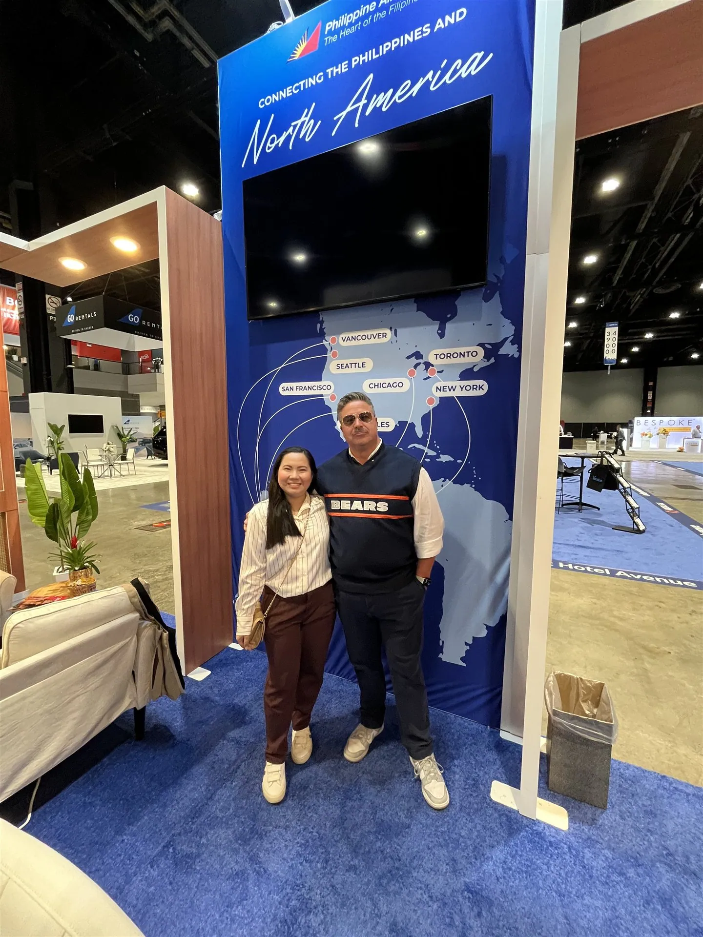 Two people posing beside Philippine Airlines’ tall blue North America route-map display and screen.