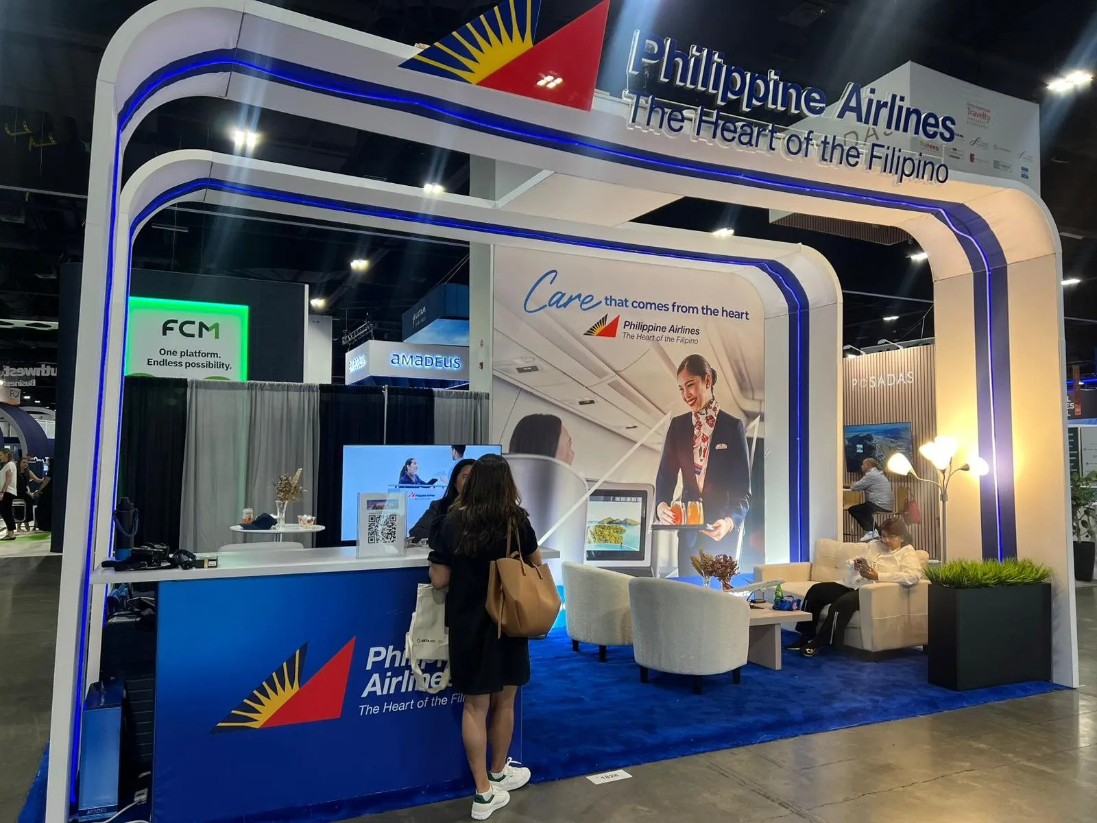 Visitor at a Philippine Airlines counter beneath curved white frames with blue lighting, with lounge seating inside the exhibit.