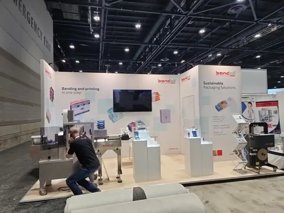 Bandall’s white exhibit with banding machines, sustainable-packaging graphics, a dark screen and a technician crouching beside equipment.