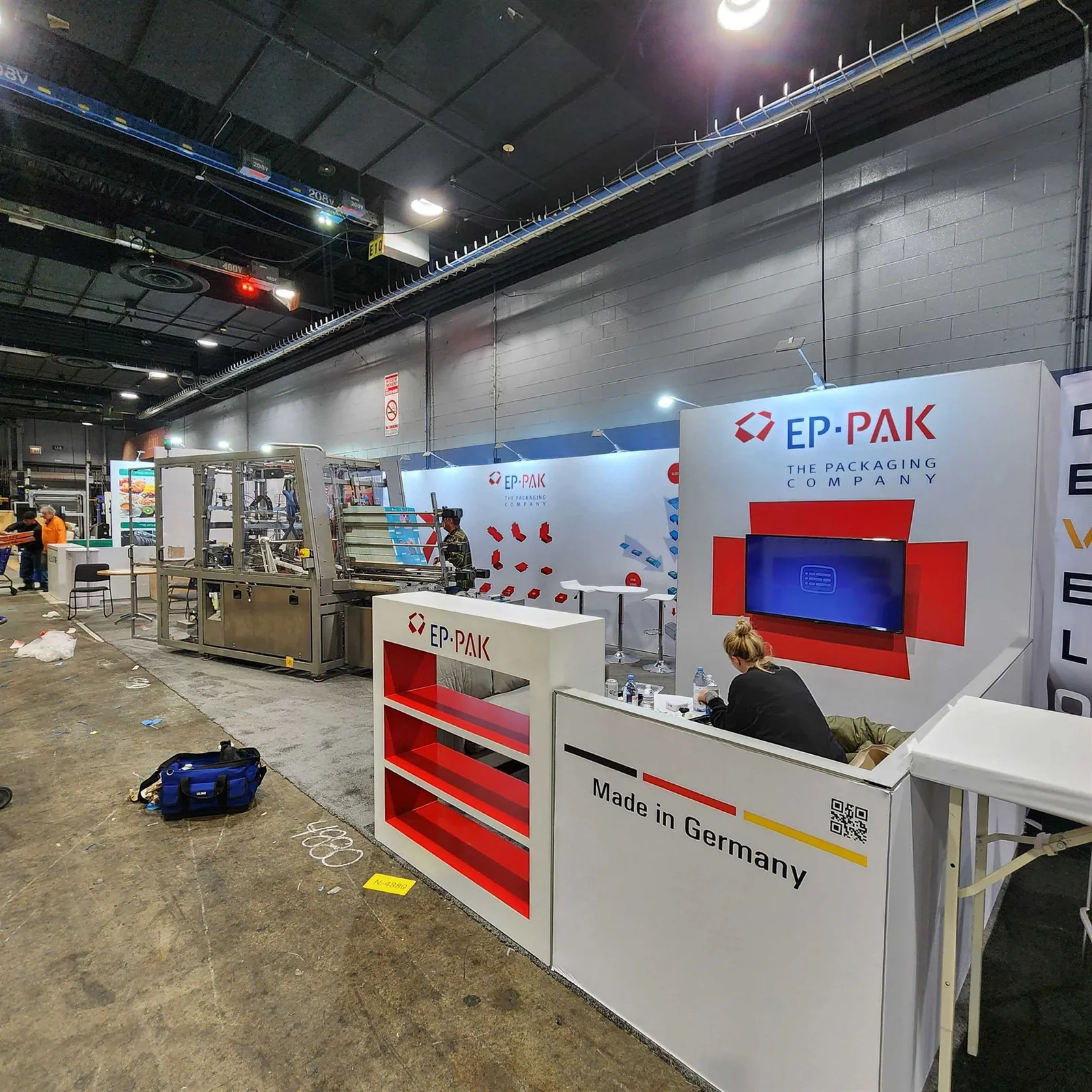 EP-PAK exhibit with a red-and-white reception counter, wall-mounted screen and enclosed packaging machinery along the left side.