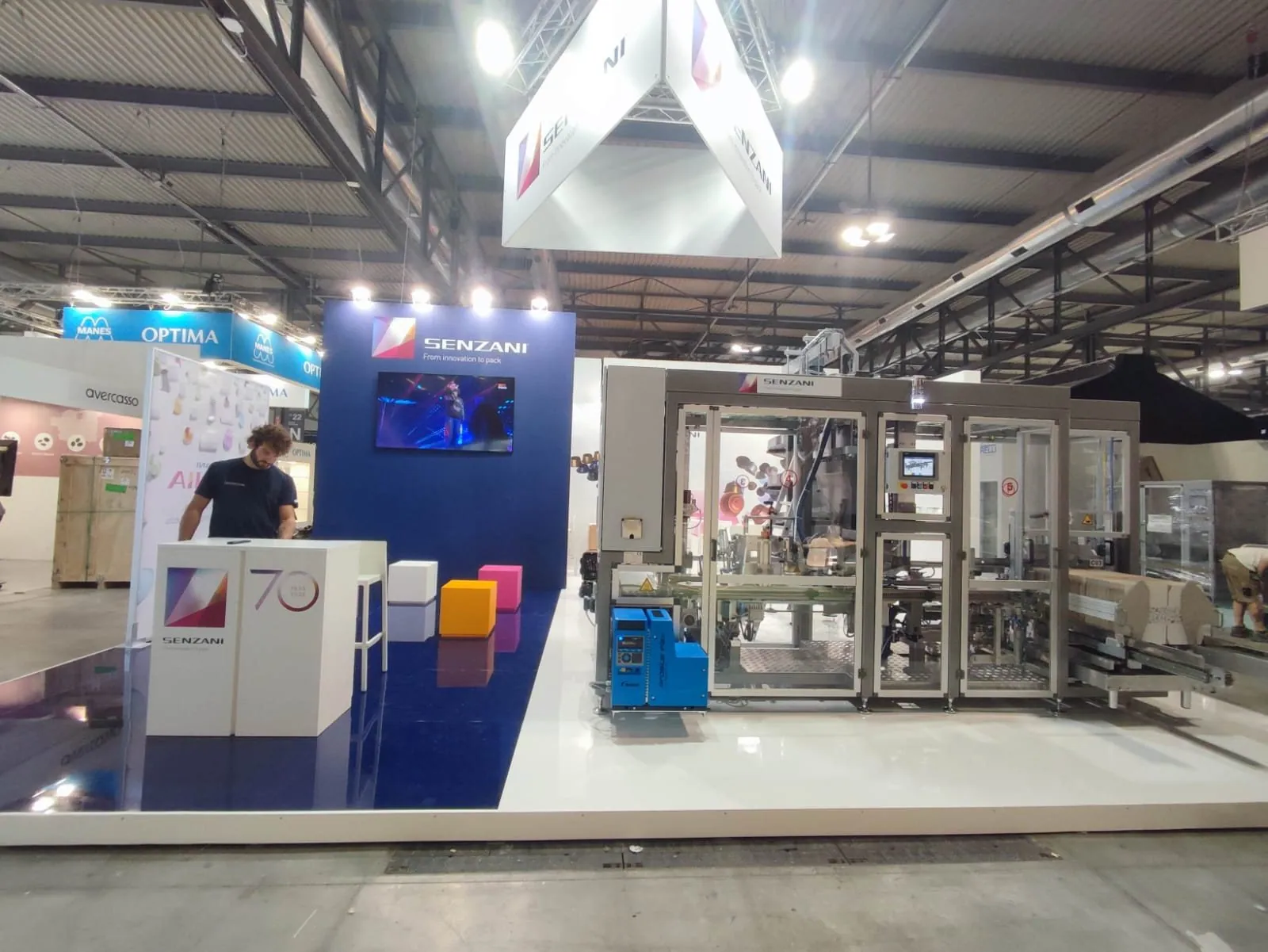 Senzani exhibit with a large enclosed packaging machine, a control screen, purple branded wall, suspended cube sign and a person at the reception counter.