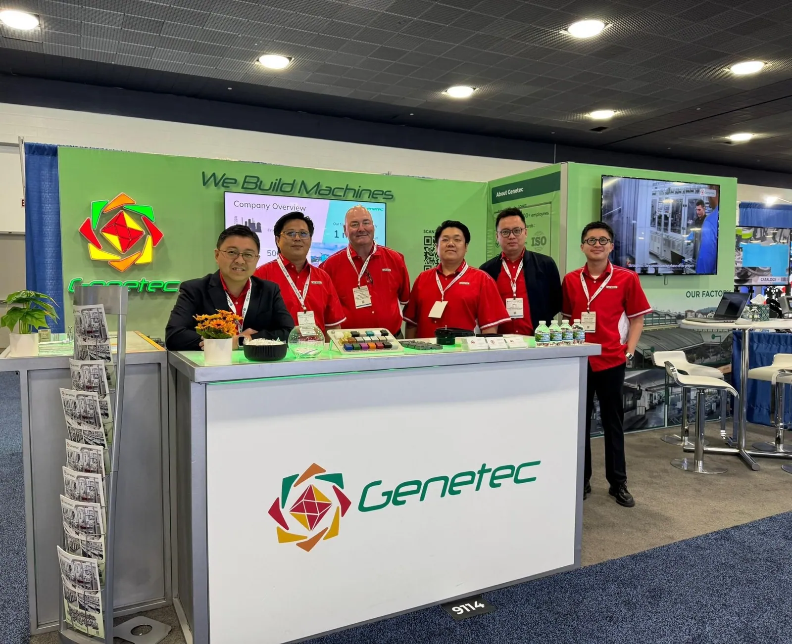 Genetec Technology team behind a white branded reception counter in a green exhibit with machine displays and factory videos.