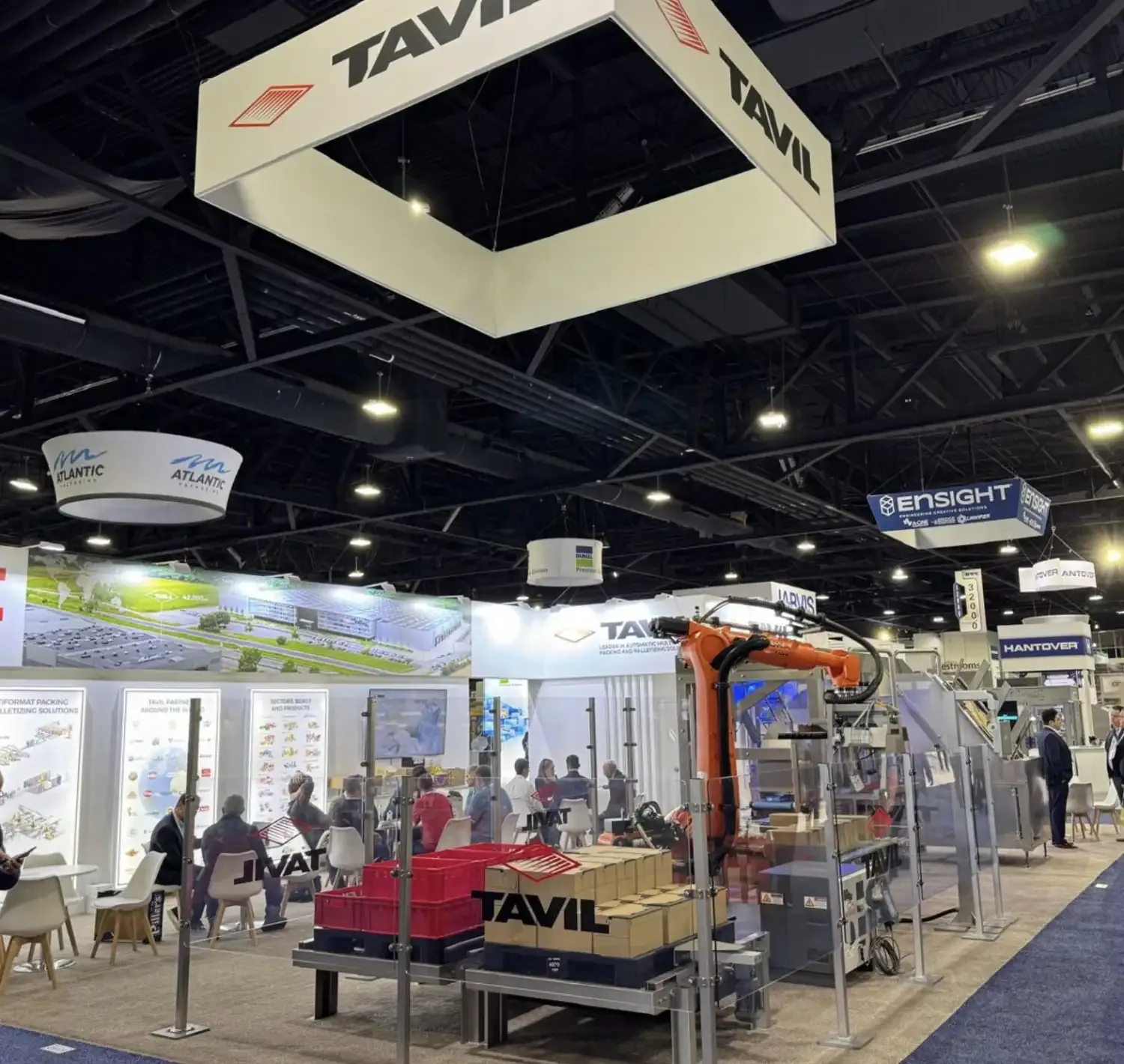 TAVIL exhibit with a suspended square sign, an orange industrial robot inside a guarded cell and visitors seated at nearby tables.
