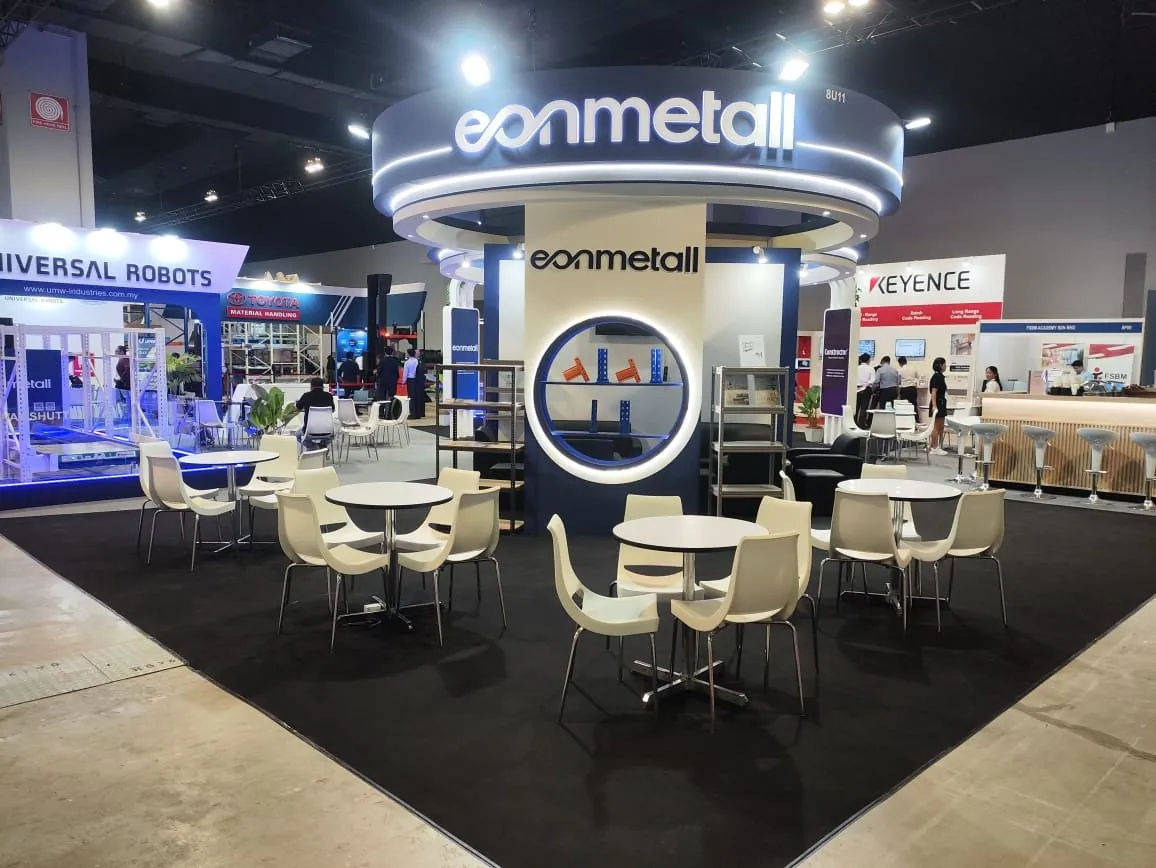 Eonmetall exhibit with an illuminated circular overhead sign, a central branded display, round meeting tables and white chairs.