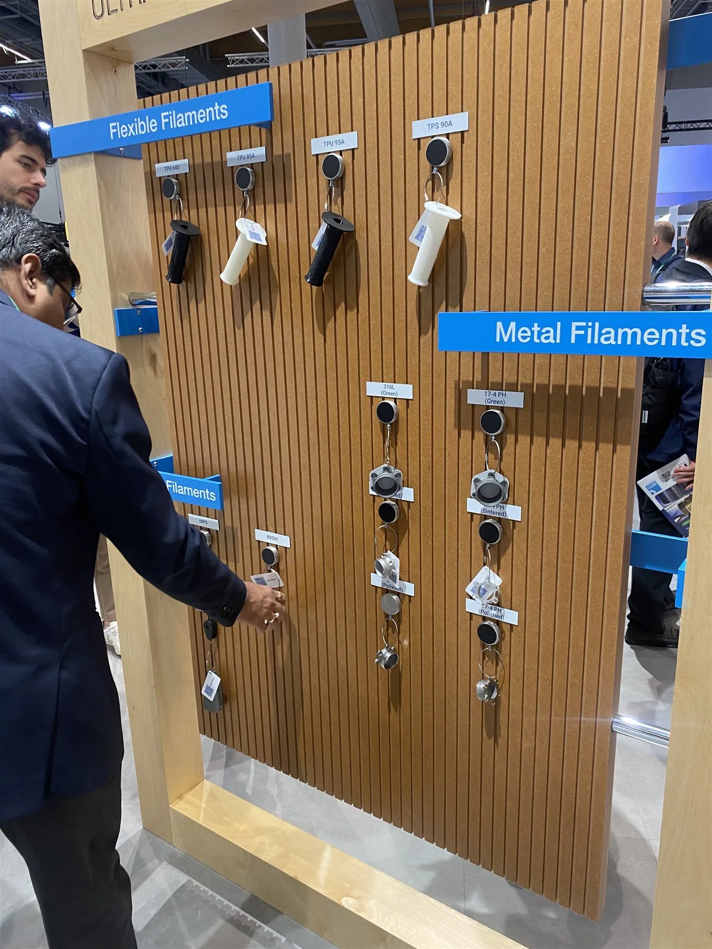 A visitor handling a tethered sample on a timber slat display labeled Flexible Filaments and Metal Filaments.