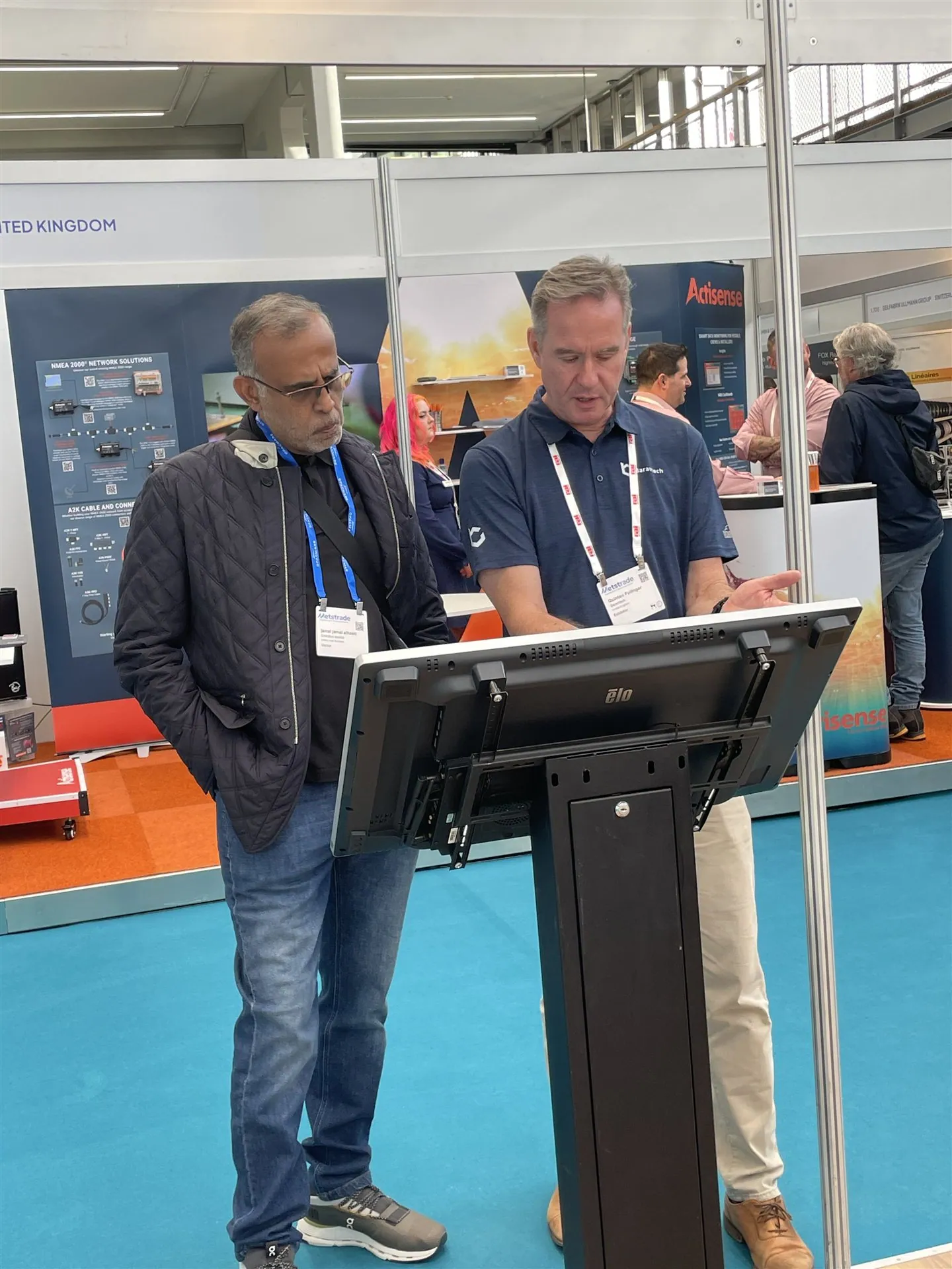 Two people discussing a large touchscreen in a marine-electronics exhibit, with Actisense network-product displays behind them.