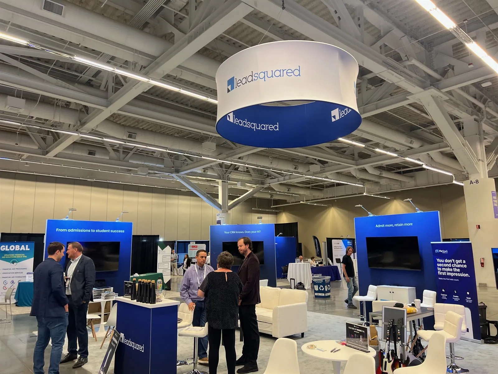 Visitors talking at LeadSquared's blue-and-white software exhibit beneath a large circular sign, with screens and consultation seating.