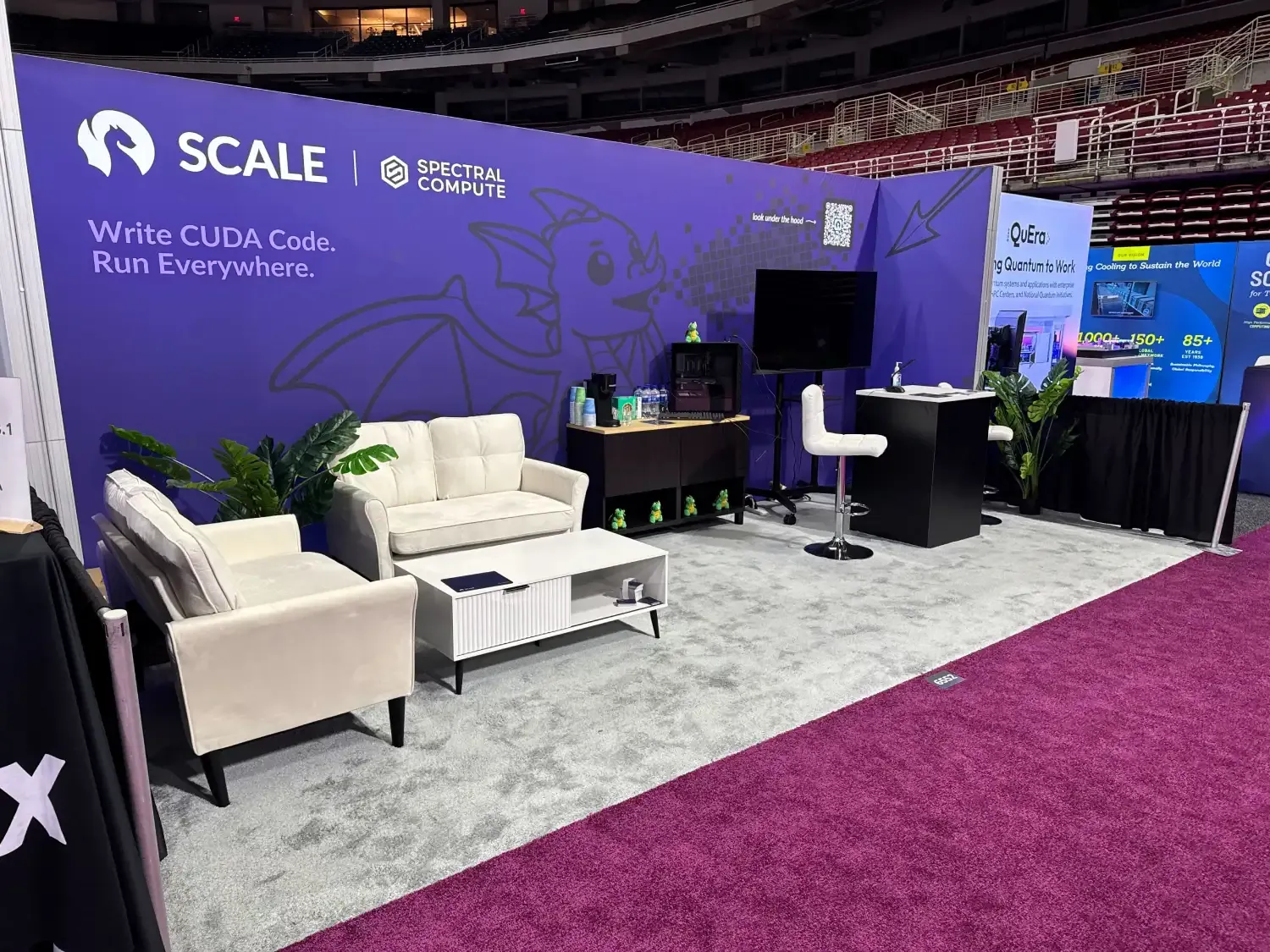 Angled view of Spectral Compute's purple exhibit with white sofas, software messaging, a monitor and a presentation counter.