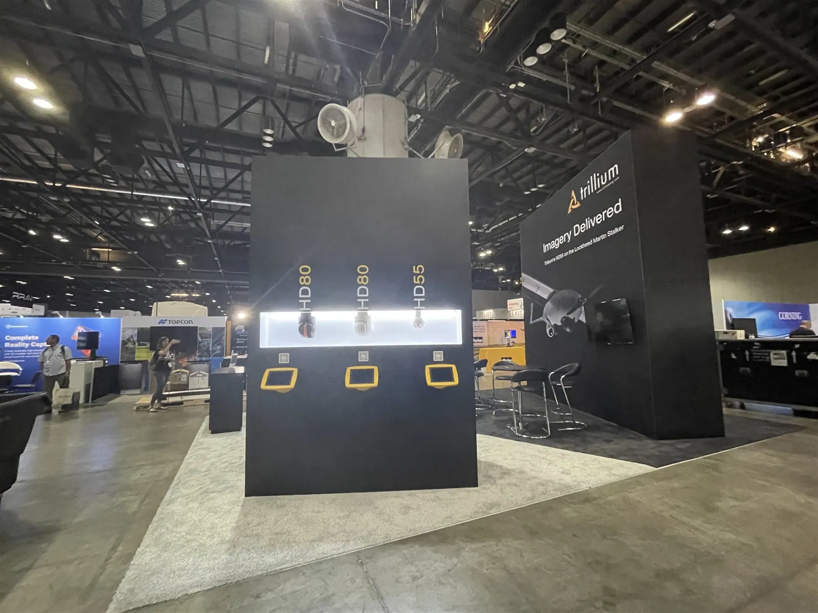 Trillium Engineering exhibit with illuminated imaging-sensor displays, yellow-framed screens and a dark aircraft graphic wall.