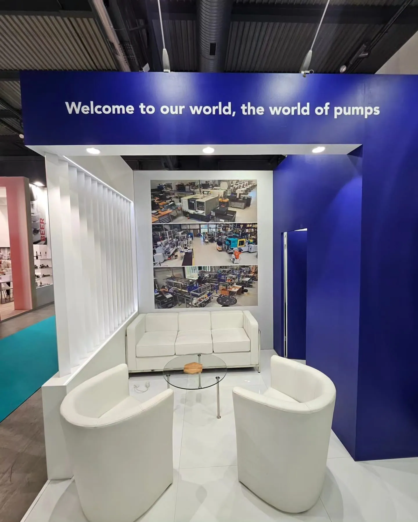 White lounge seating and a glass table beneath a blue exhibit arch reading Welcome to our world, the world of pumps.