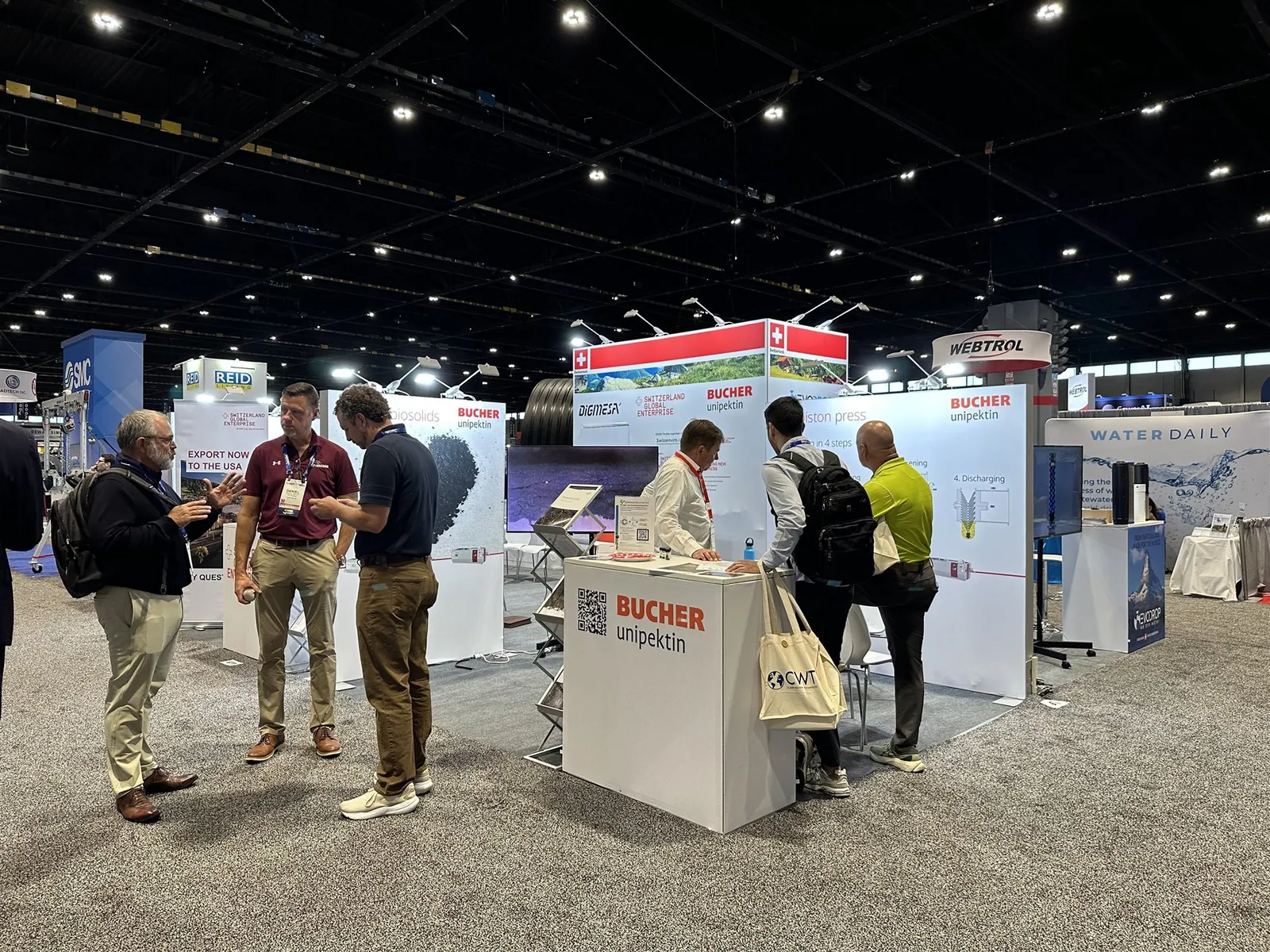 Visitors talking at the Bucher Unipektin counter in a Swiss water-technology pavilion with piston-press and biosolids graphics.