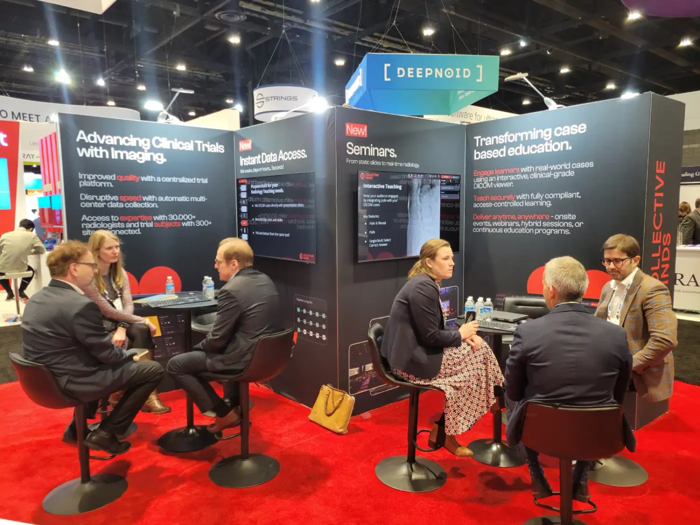 Visitors meeting at two tables in Collective Minds’ red-carpeted exhibit, surrounded by screens and wall graphics about clinical-trial imaging, data access and case-based education.