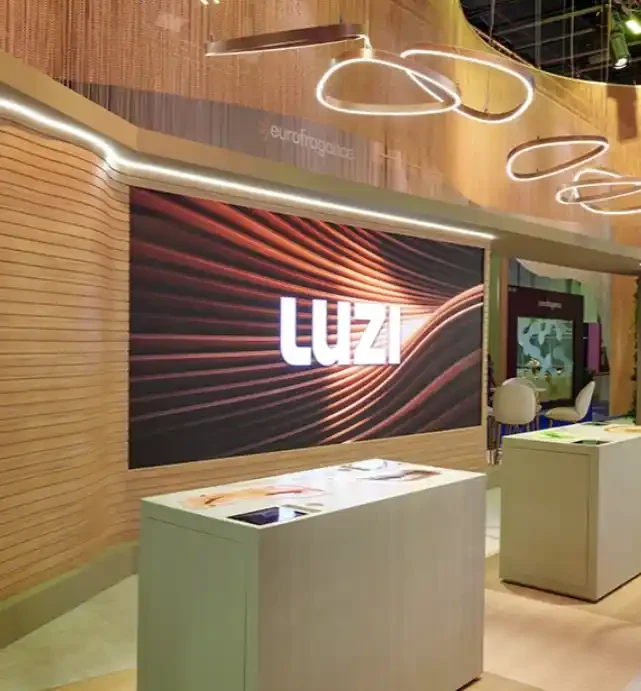 LUZI fragrance exhibit with warm wood walls, circular suspended lights, a large branded screen and sample counters.