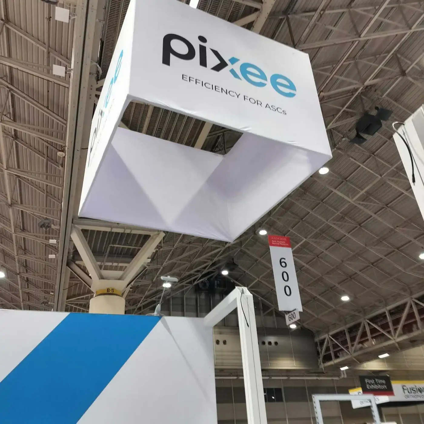 Overhead branding detail of the suspended white Pixee cube sign above the blue-and-white medical technology exhibit.