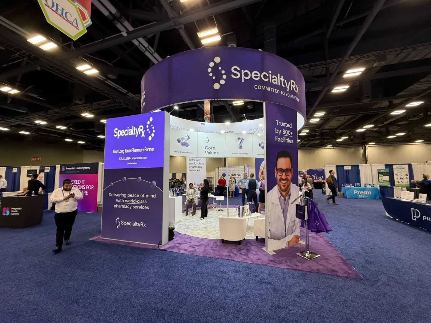 SpecialtyRx pharmacy exhibit with circular purple overhead branding, tall display pillars, seating and visitors.