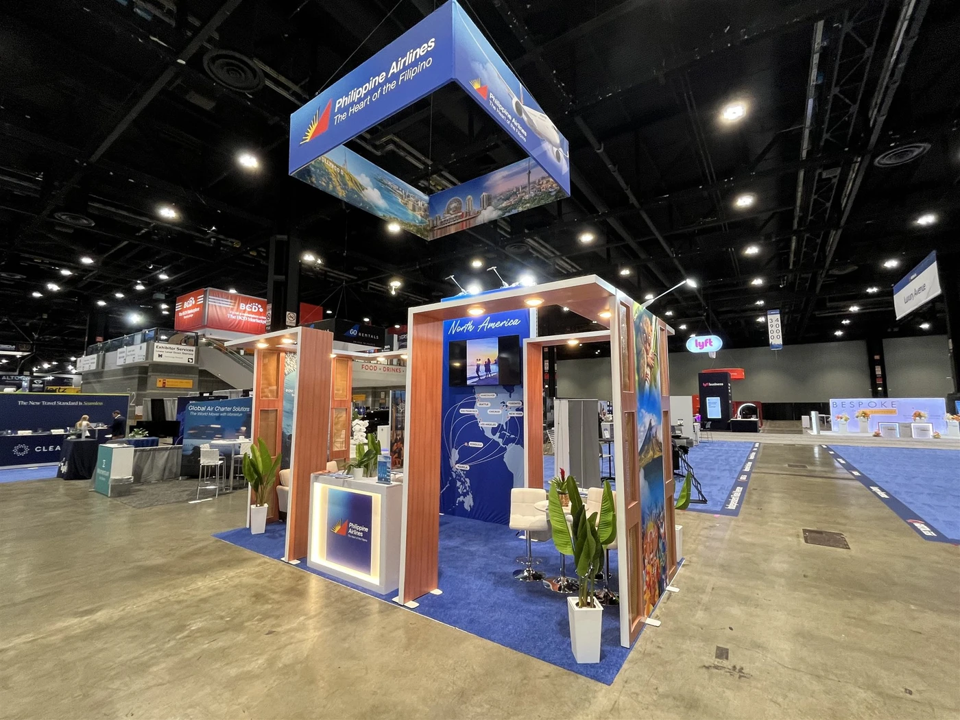 Philippine Airlines exhibit with suspended blue square branding, illuminated timber-framed portals, route graphics, a reception counter and white meeting chairs.