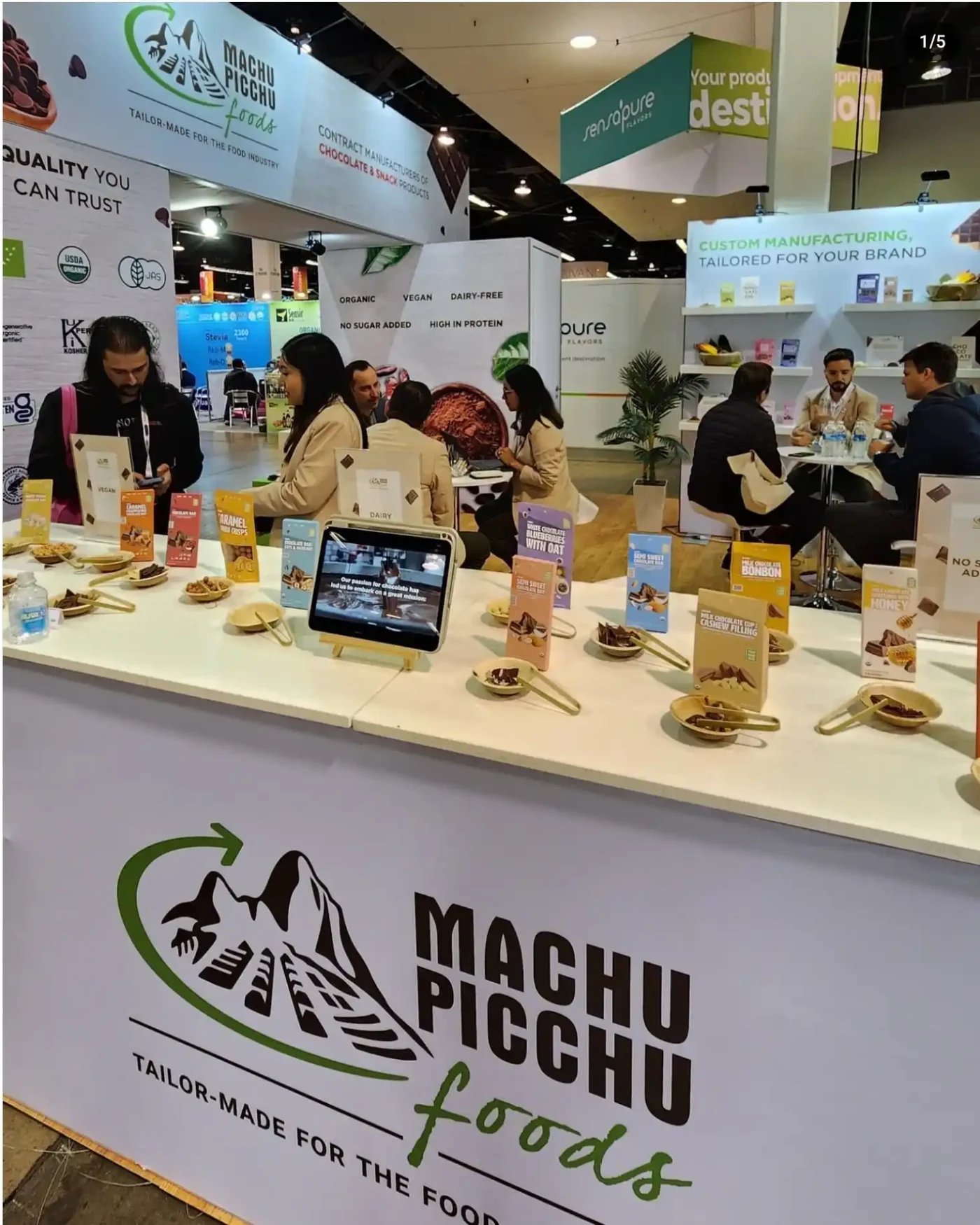 Machu Picchu Foods exhibit with a branded sampling counter, packaged food displays and visitors meeting inside the booth.