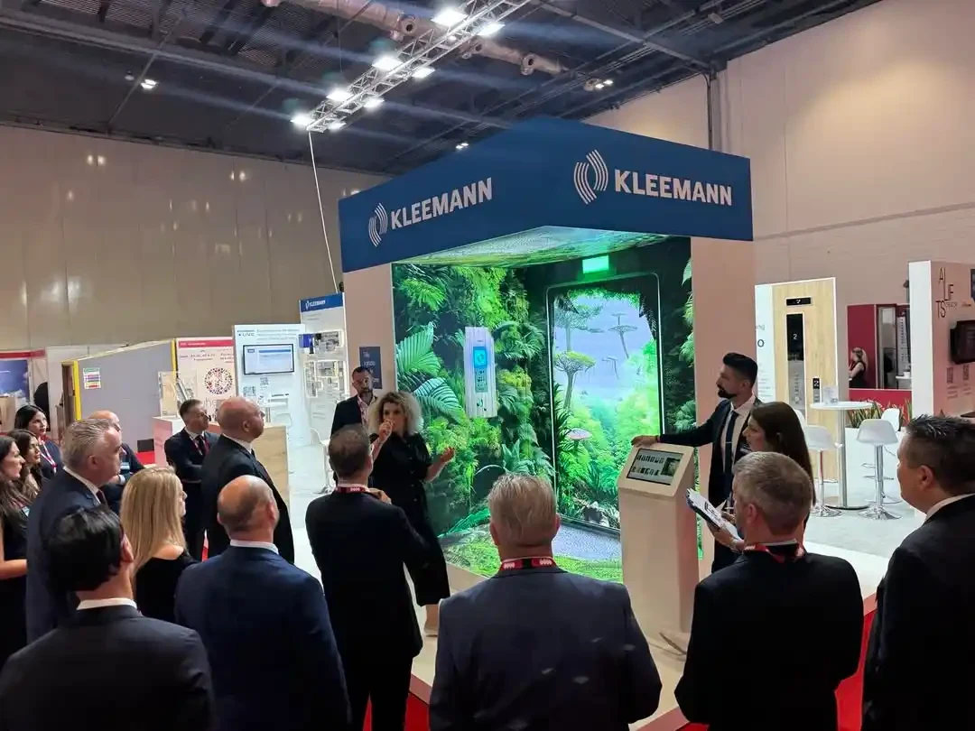 Visitors watching an elevator technology presentation beneath KLEEMANN branding, with an illuminated forest-themed display and a touchscreen kiosk.