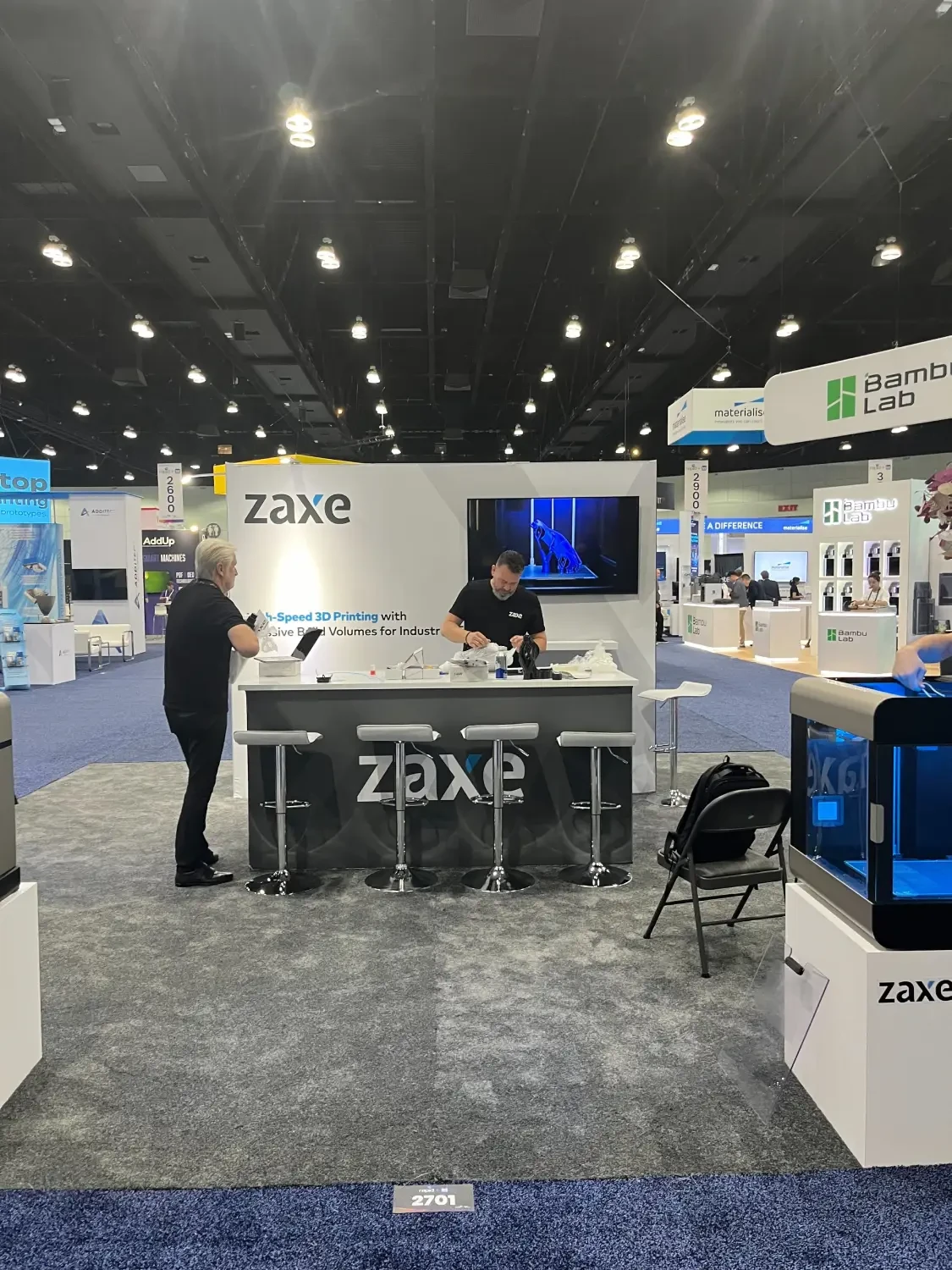 Zaxe exhibit with a blue-lit enclosed 3D printer in the foreground, a branded sample counter, two people and a wall-mounted product screen.