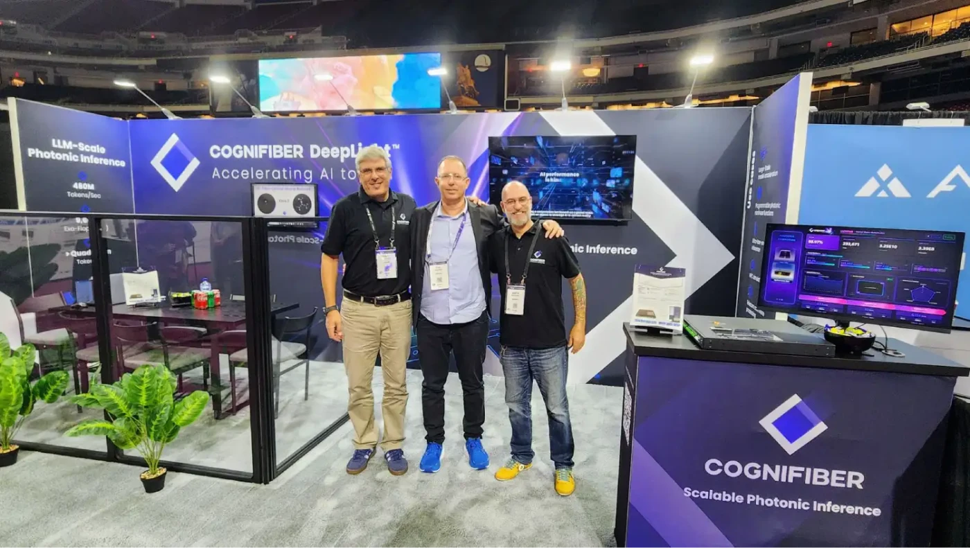 Three people standing in Cognifiber’s purple-and-white exhibit beside a photonic-computing demonstration counter, performance monitor and glass meeting area.