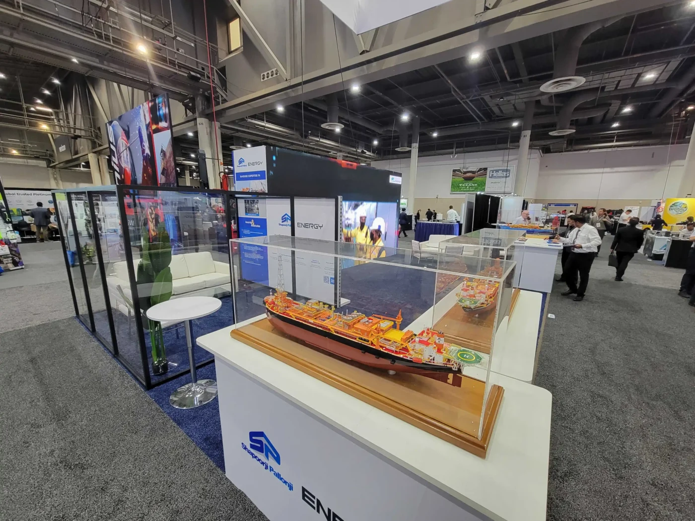 An offshore production vessel model in a clear display case beside Shapoorji Pallonji Energy's glass meeting room, branded screens and blue carpet.