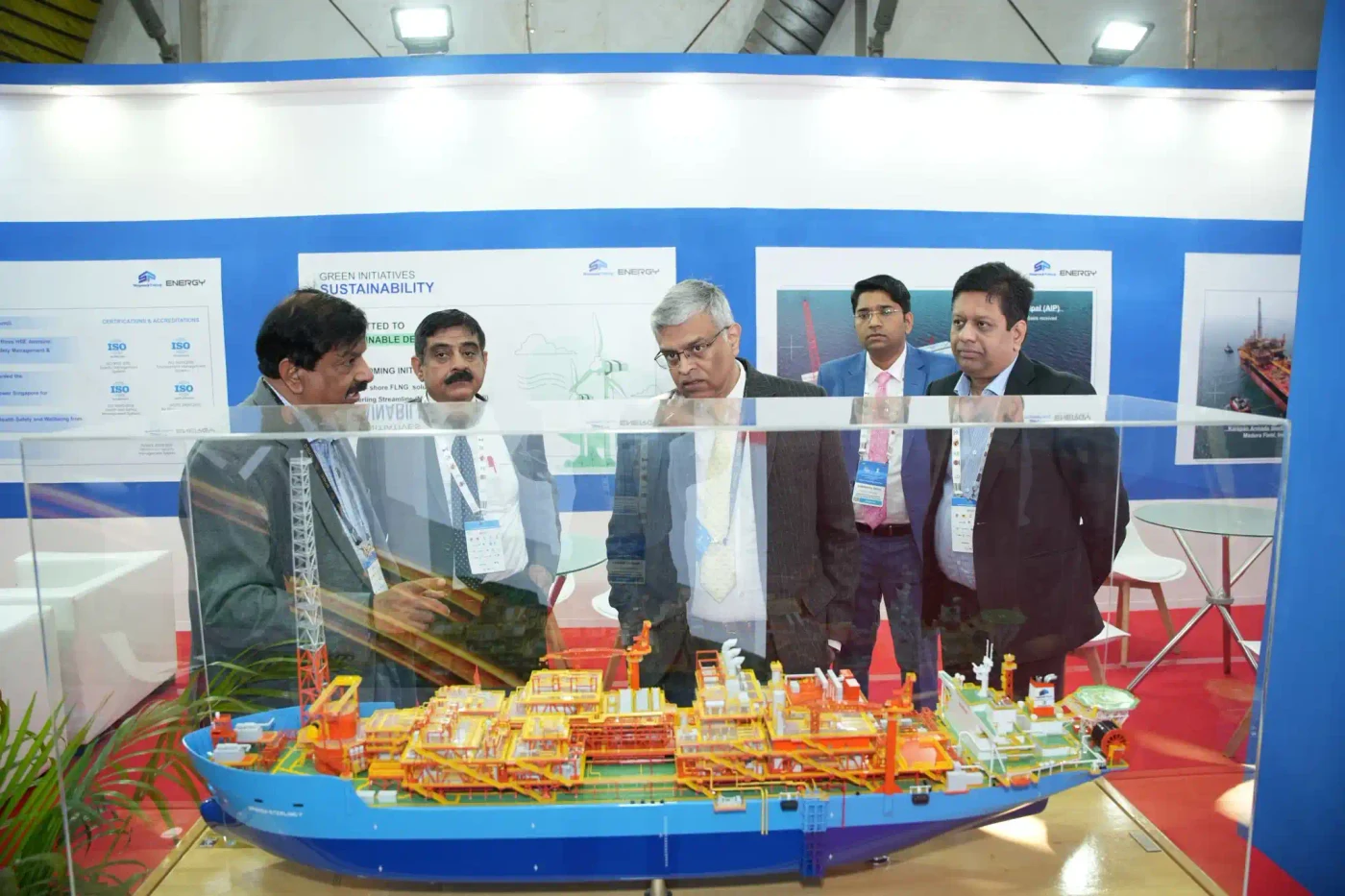Visitors discuss a detailed offshore vessel model inside Shapoorji Pallonji Energy's blue-and-white exhibit, with project information on the wall behind them.