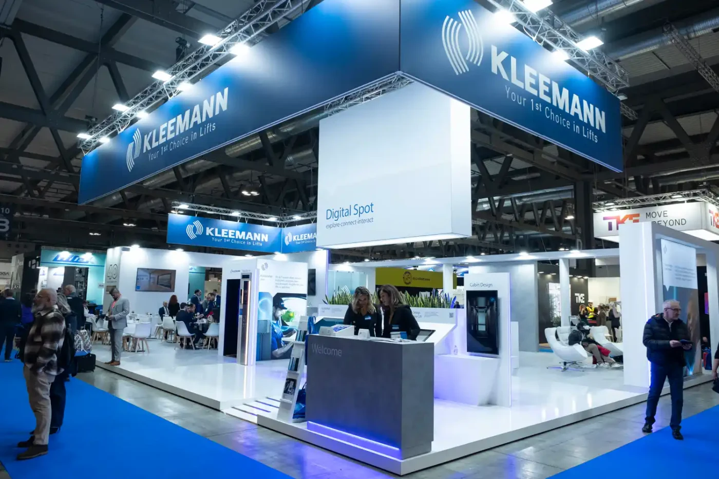 KLEEMANN elevator exhibit with large suspended blue signage, a Digital Spot demonstration area, product displays and meeting tables.