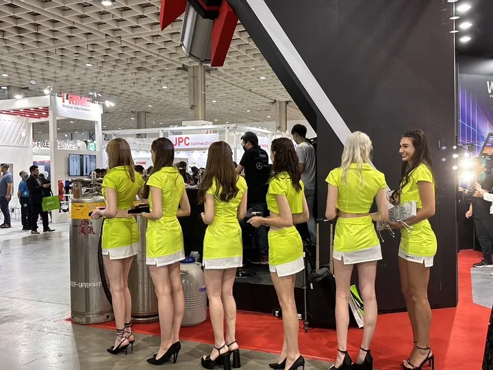 promotional models working at a technology exhibit booth with attendees and surrounding trade show booths visible.