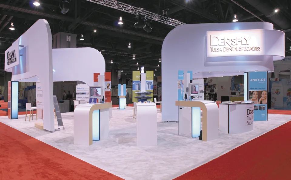 Dentsply Tulsa Dental Specialties 30x30 island exhibit with curved white overhead structures, backlit demo counters and product stations