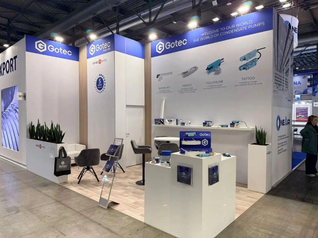 Gotec 10x20 corner inline trade show booth with white walls, blue headers, product shelf and a seated meeting area